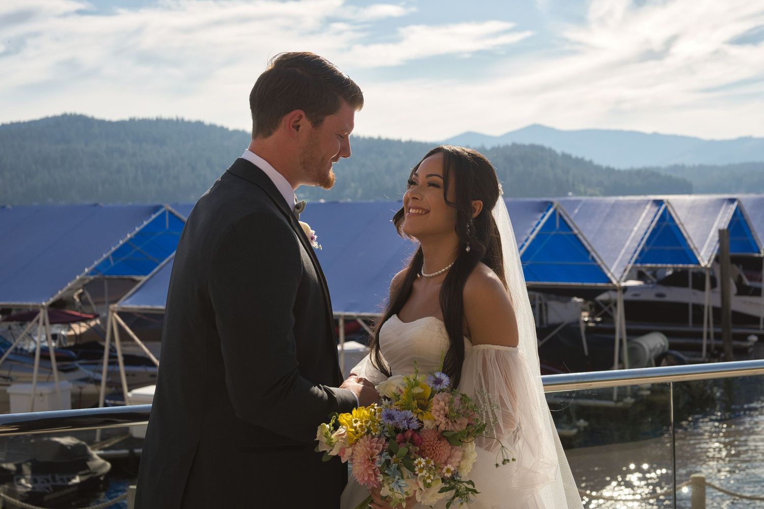 Wedding couple smile as they look at each other, standing outside in front of Lake Coeur d' Alene.