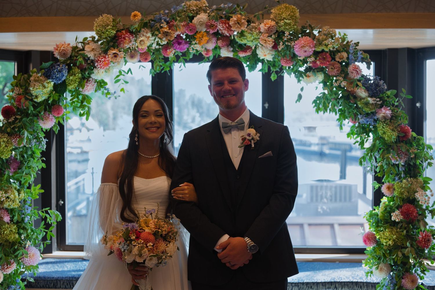 Bride and groom pose for a formal portrait in front of their arch, with colorful flowers and sunlight around them.