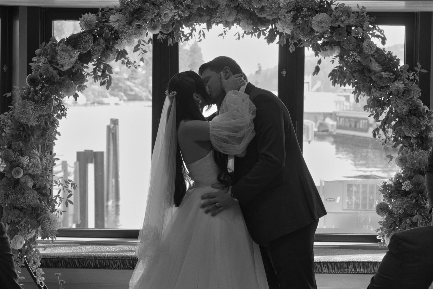 Black and white portrait of the couple kissing, standing in front of their flower arch during the ceremony.