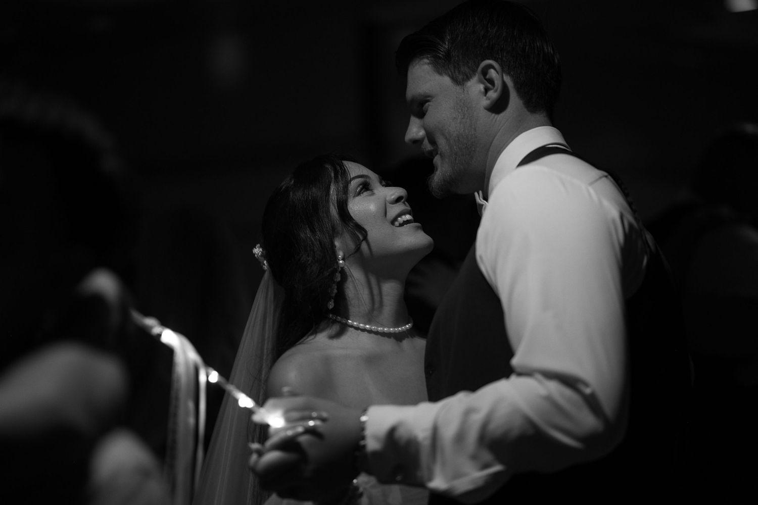 Black and white photo of bride looking up at groom while they dance together under a light creating dramatic shadows.