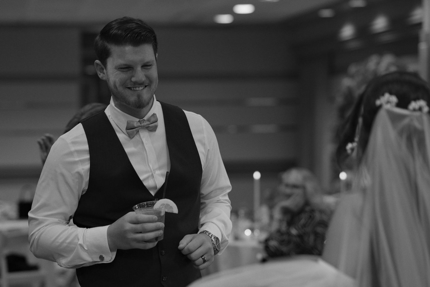 Black and white picture that focuses on the groom as he smiles at his bride, out of focus closer to the camera.