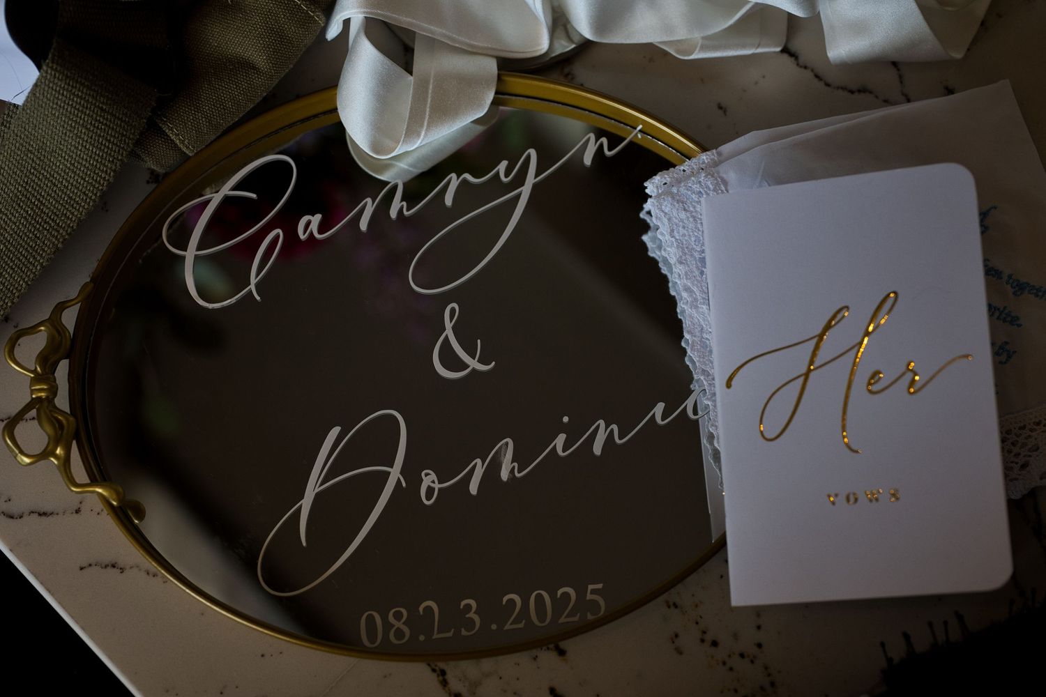 Flat-lay photo of the bride's vow book, ribbon, and a mirror with the bride and groom's names painted across.