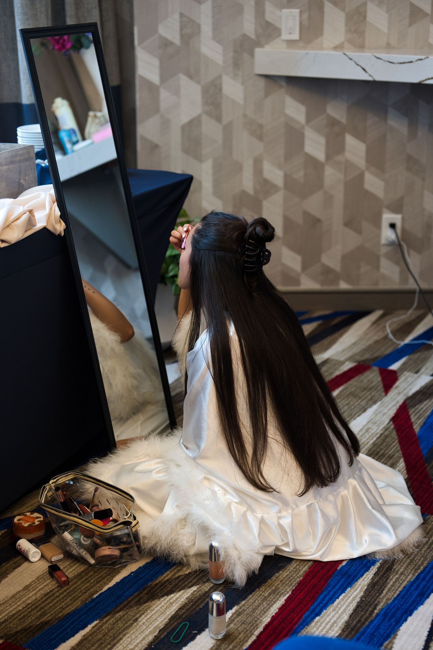 Bride sits cross-legged on the floor in front of a mirror as she gets ready, putting on her makeup.