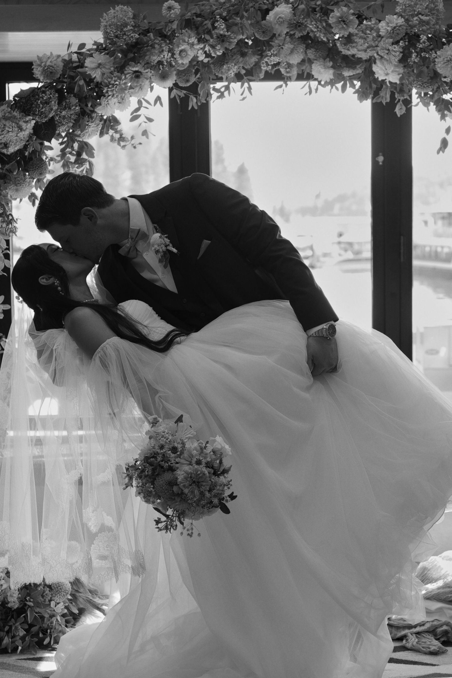 Black and white picture of the bride and groom kissing as he dips her in front of the wedding arch covered in flowers.