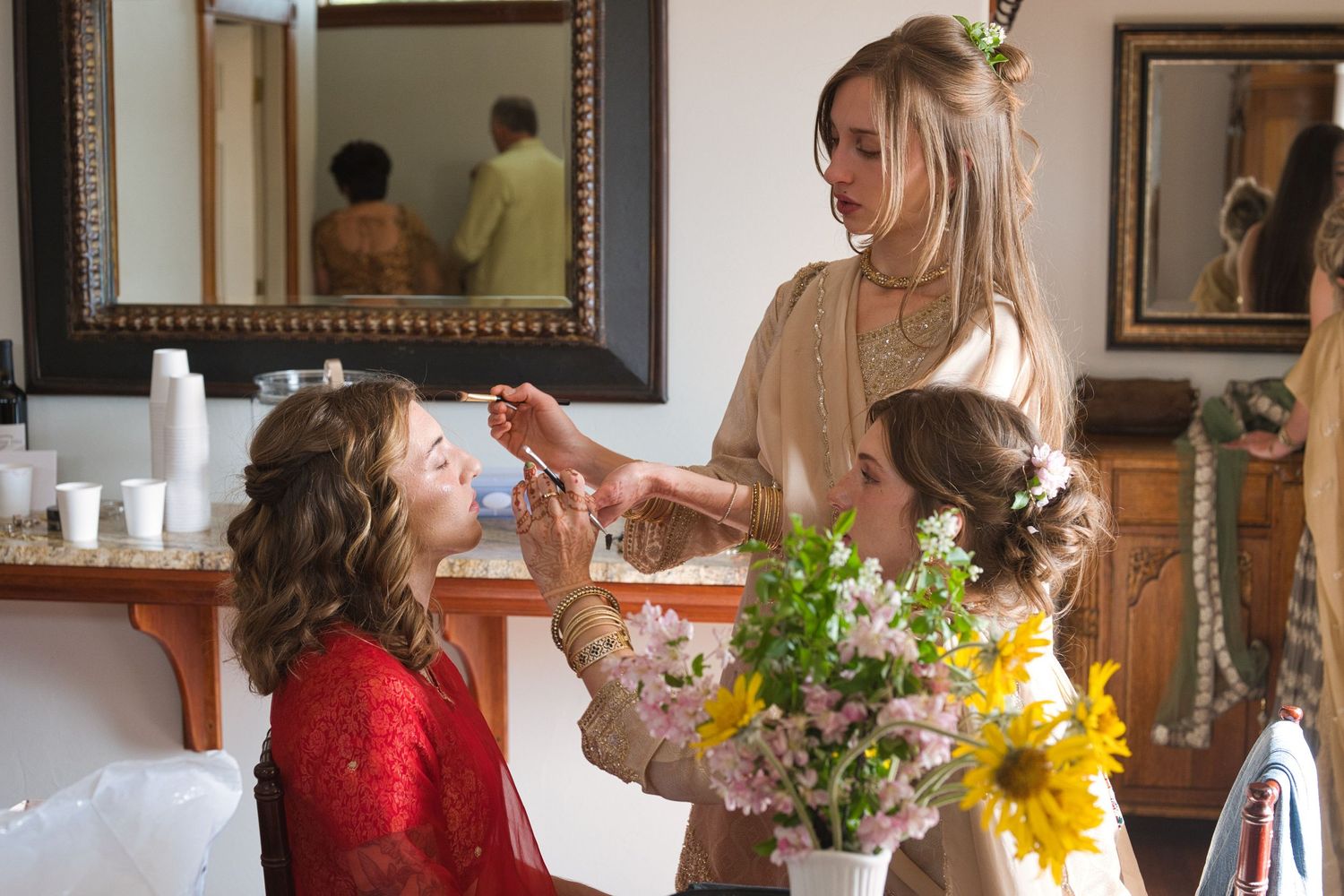 Bride sits while two bridesmaids apply her makeup with brushes in the bridal suite, natural light shining in.