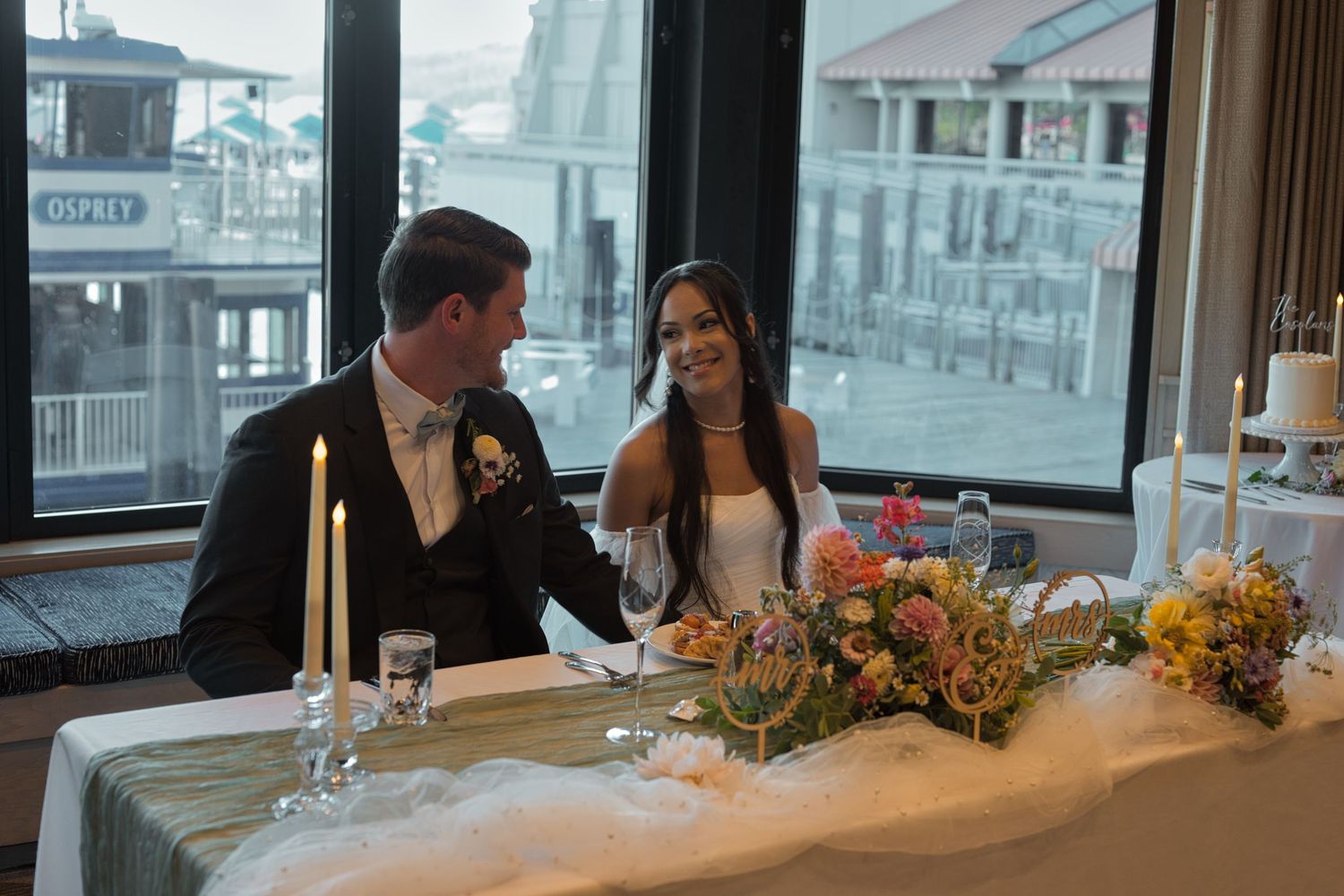 Bride and groom smile at each other as they sit down at the table in front of large bay windows, looking out over Coeur d' Alene Lake.