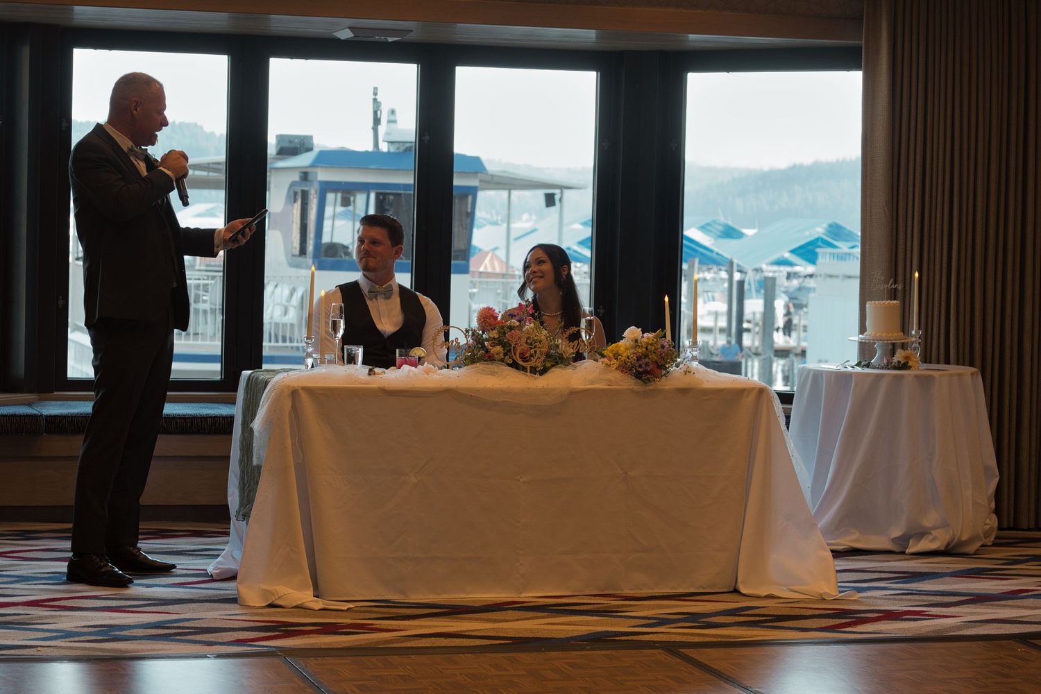 Bride and groom look at the bride's father as he toasts to them during a speech, he is holding a microphone.