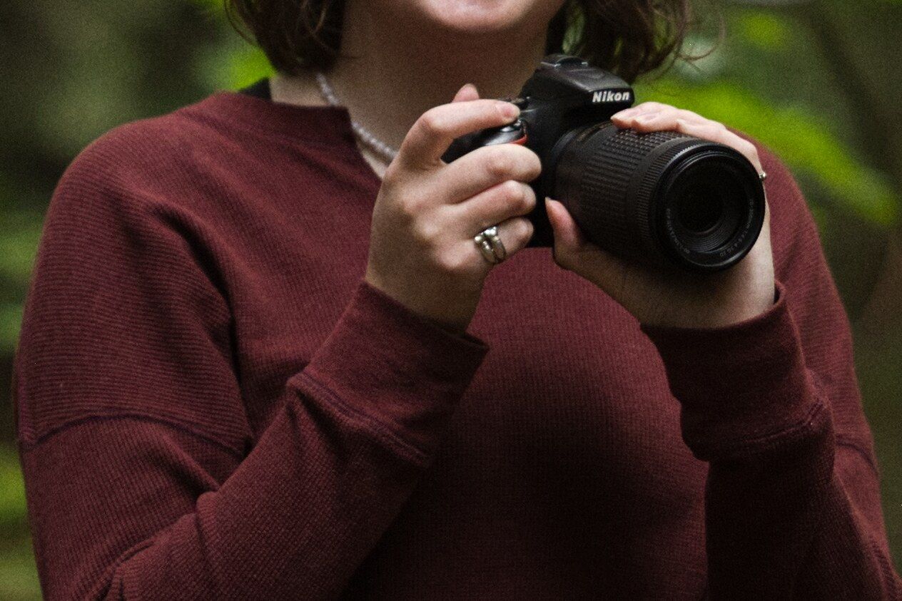 A close up picture focusing on the hands of a girl in a red shirt holding a Nikon camera in Montana forest.