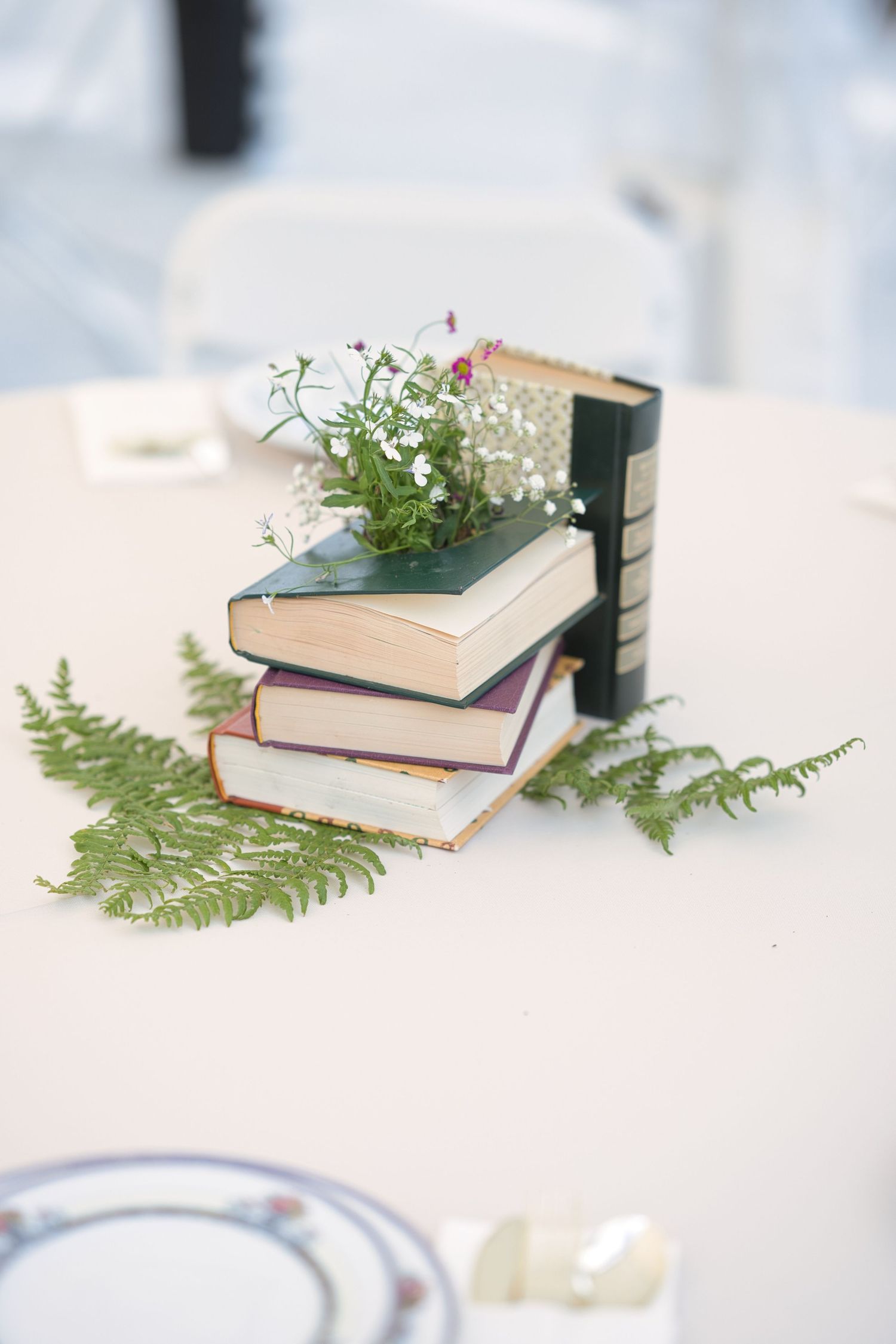 Table decoration of stacked books and flowers bursting out of the top, with fern fronds placed below in North Idaho Wedding.