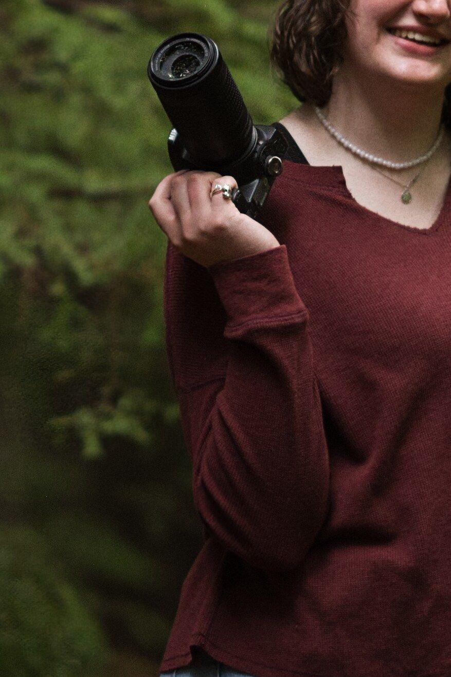Cropped picture of a girl holding a camera with her elbow on her hip as she smiles in Montana forest.