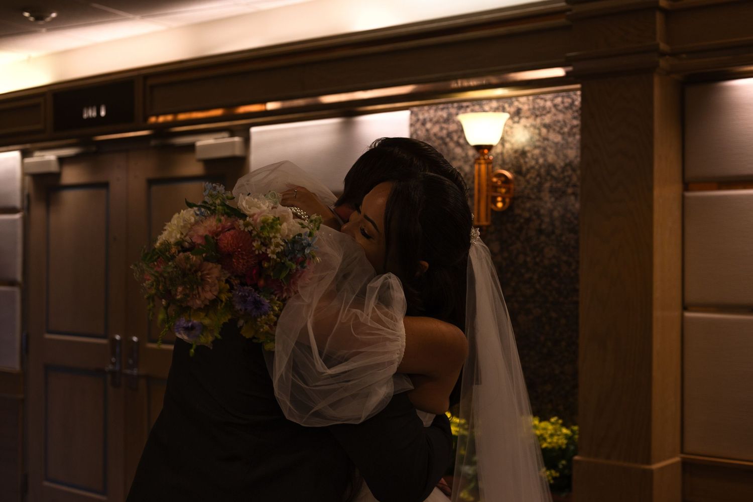 Bride and groom embrace in a tight hug immediately after leaving their wedding ceremony, standing in a large hallway.
