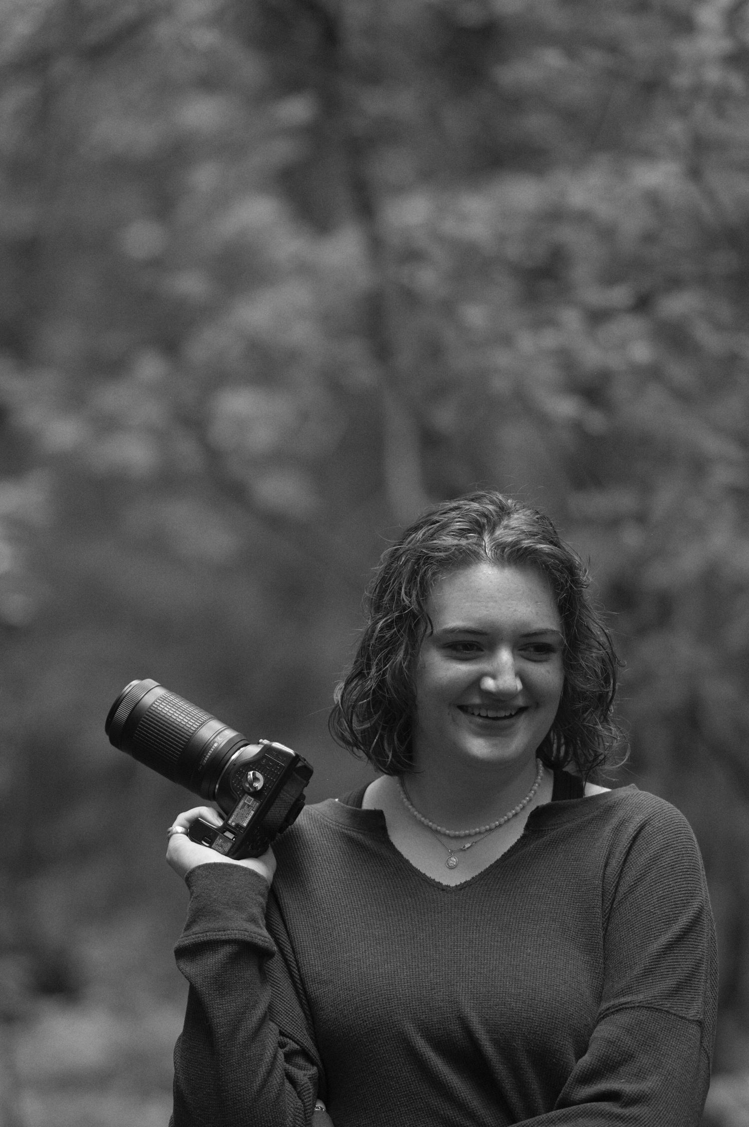 Black and white photograph of a girl standing in a Montana forest holding a camera with her elbow propped on her hip.