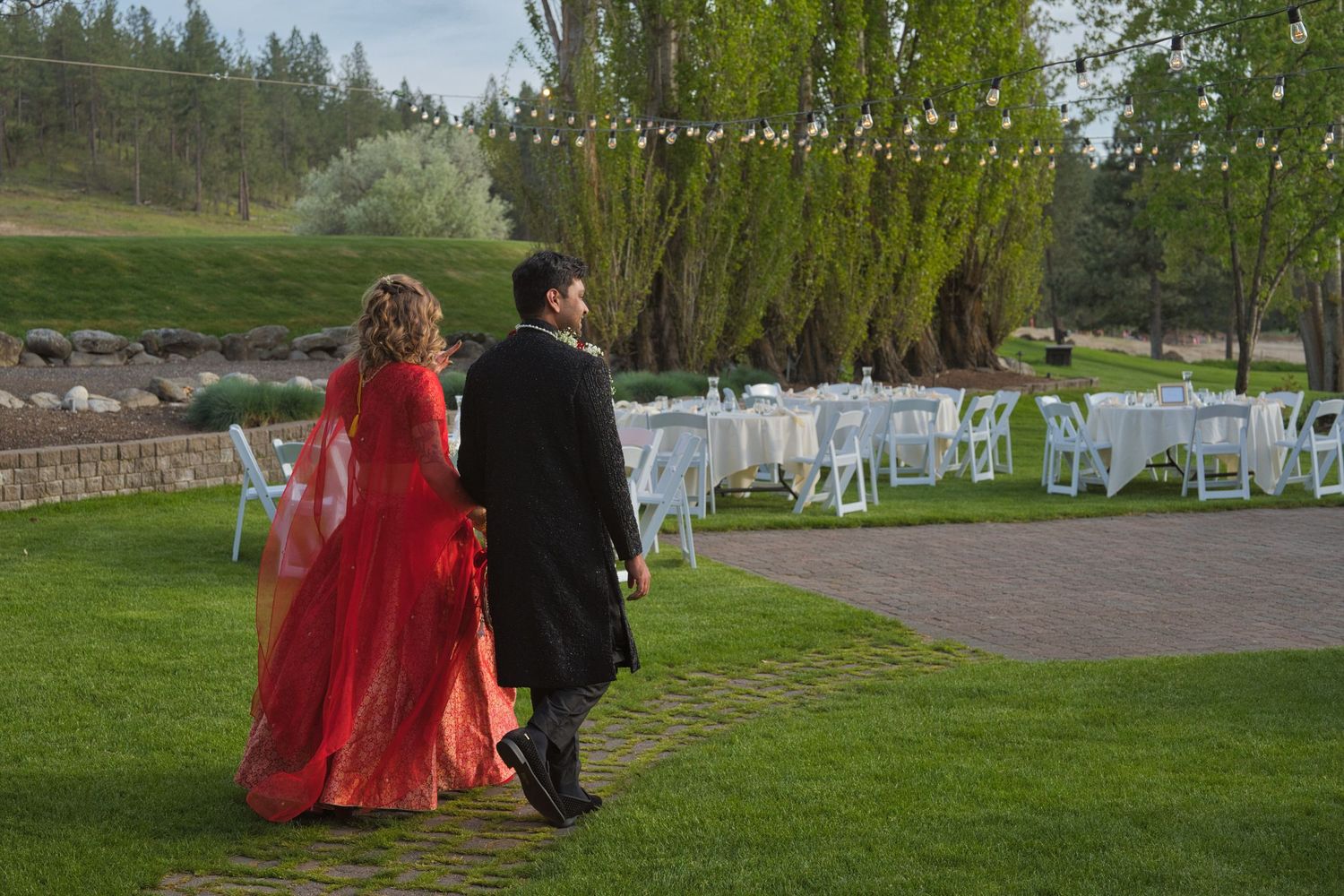 Groom leads bride to their table for the wedding reception, lights strung above and white tables in the background.