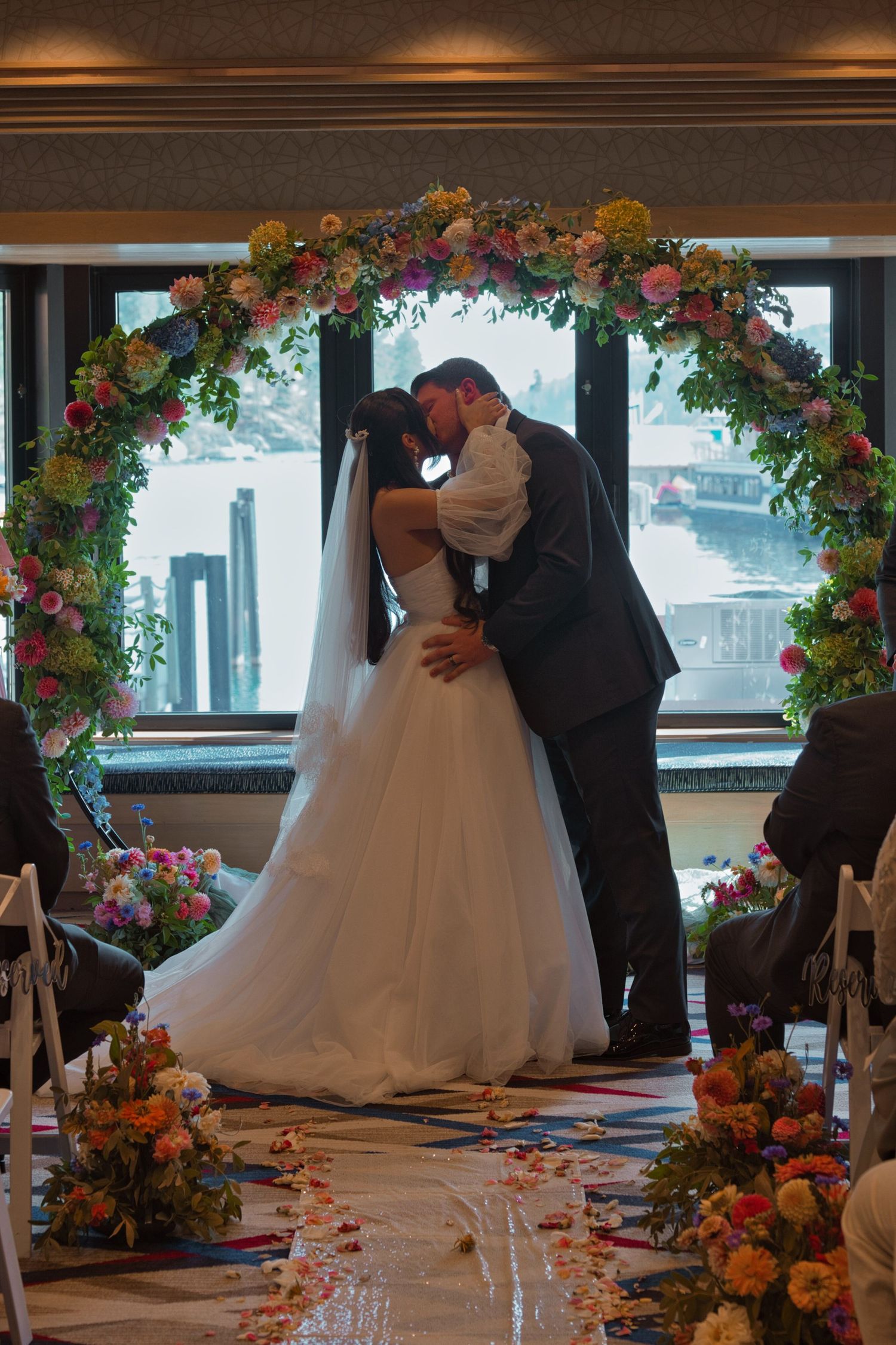 Bride and groom's first kiss during their wedding ceremony, as they stand in front of large windows, letting in the sunlight.