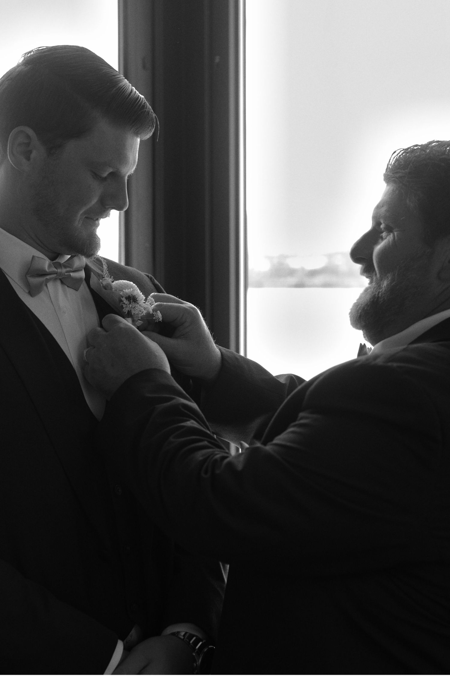 Black and white picture of the groom's father pinning the boutonniere to the groom's jacket, standing in front of a window.