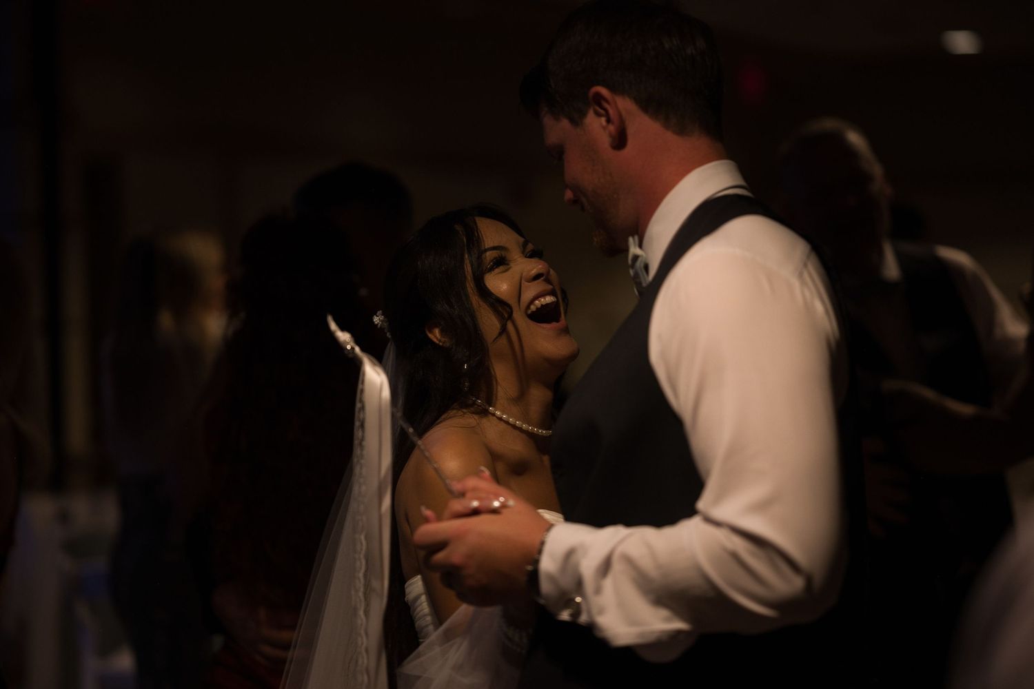 Bride laughs looking at her groom while they dance together during their reception while a light makes dramatic shadows.