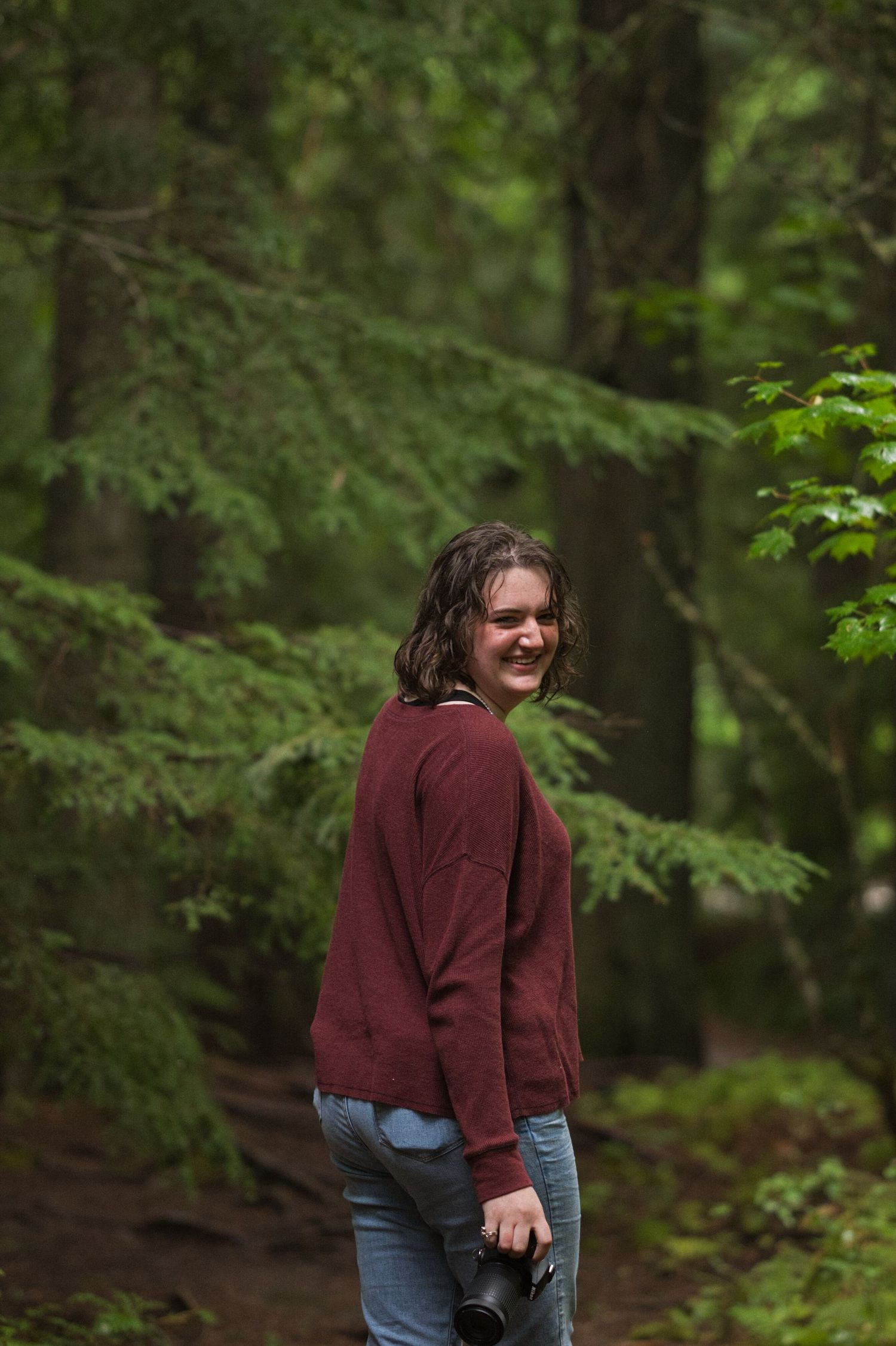 A girl laughs, looking over her shoulder while holding a camera on a walk through the dark green Montana forest.