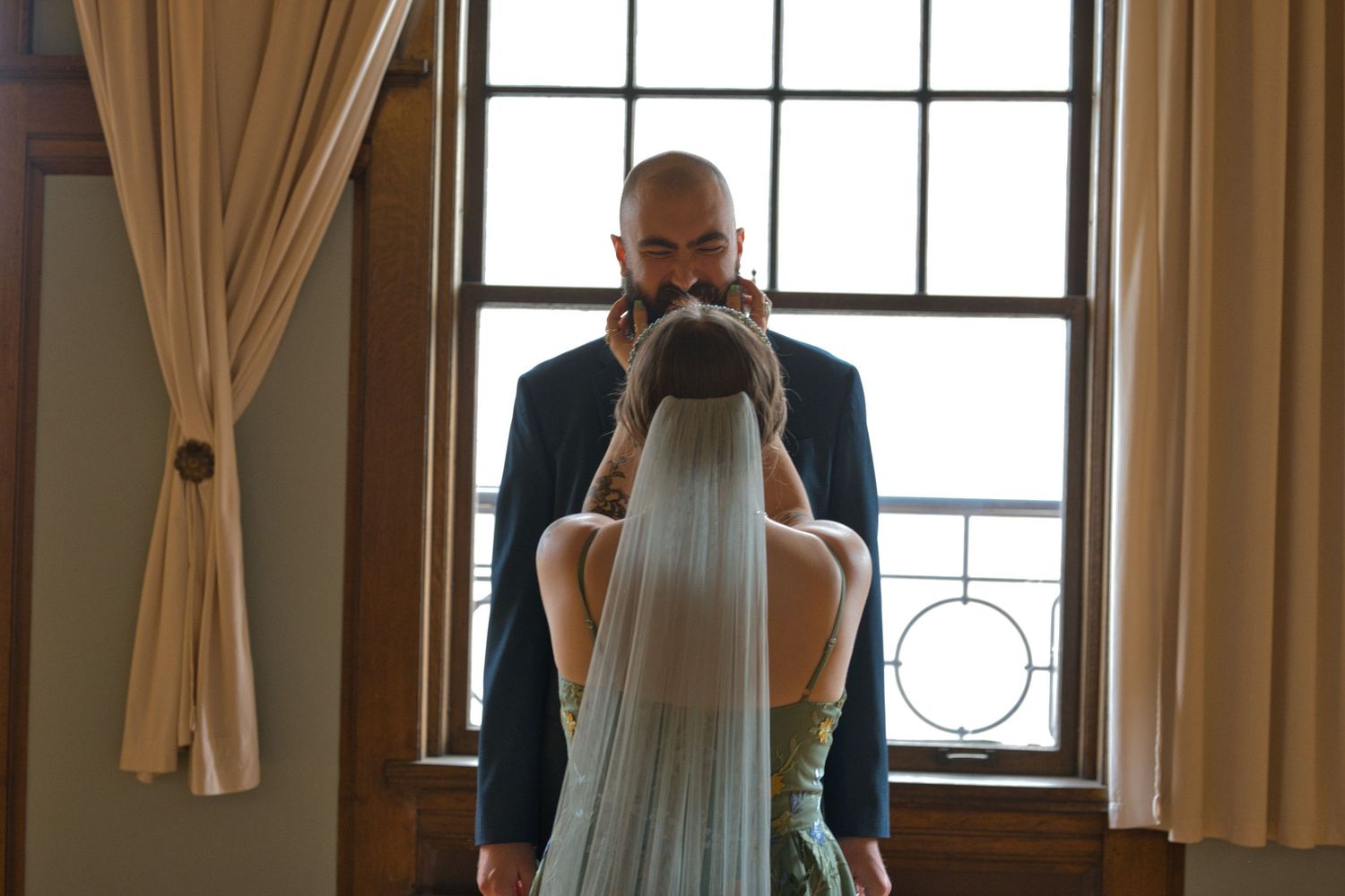 Bride reaches up to touch groom's face, a window behind them lighting up their faces.
