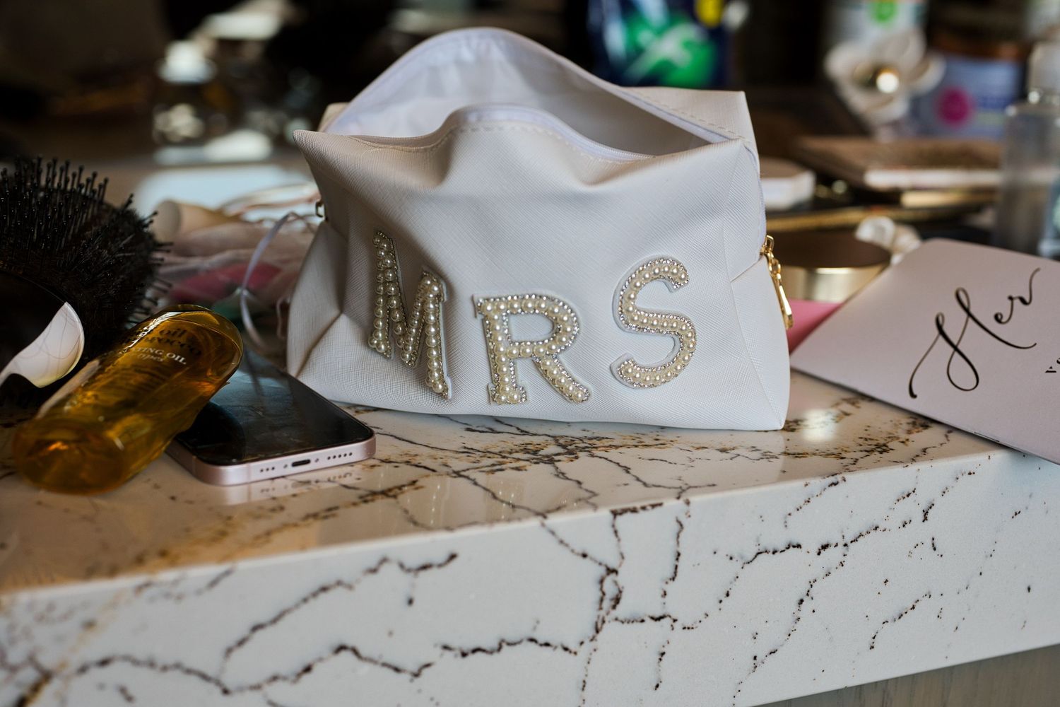 Bride's makeup bag with 'Mrs.' beading surrounded by vow book, makeup and getting ready supplies.