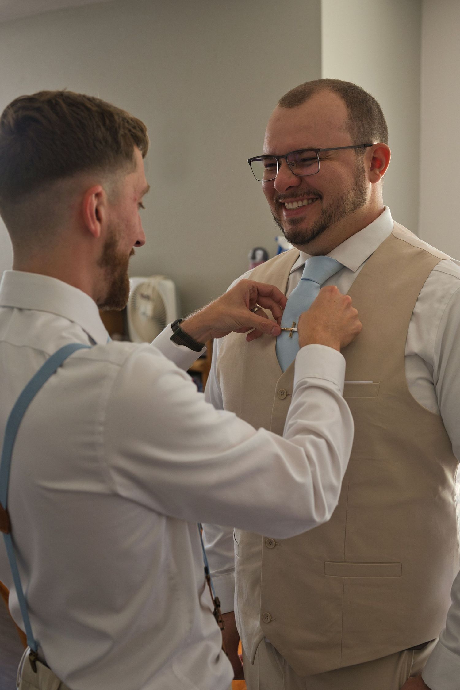 Groomsman adjusts groom's tie while both laugh