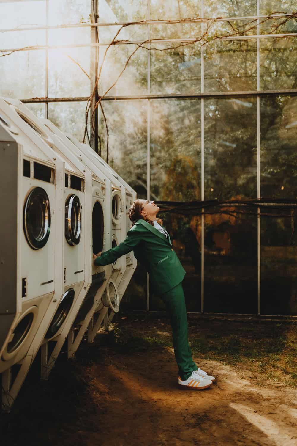 Een persoon in een groen pak leunt tegen vintage wasmachines. Zonlicht stroomt door grote ramen en creëert een nostalgische sfeer.