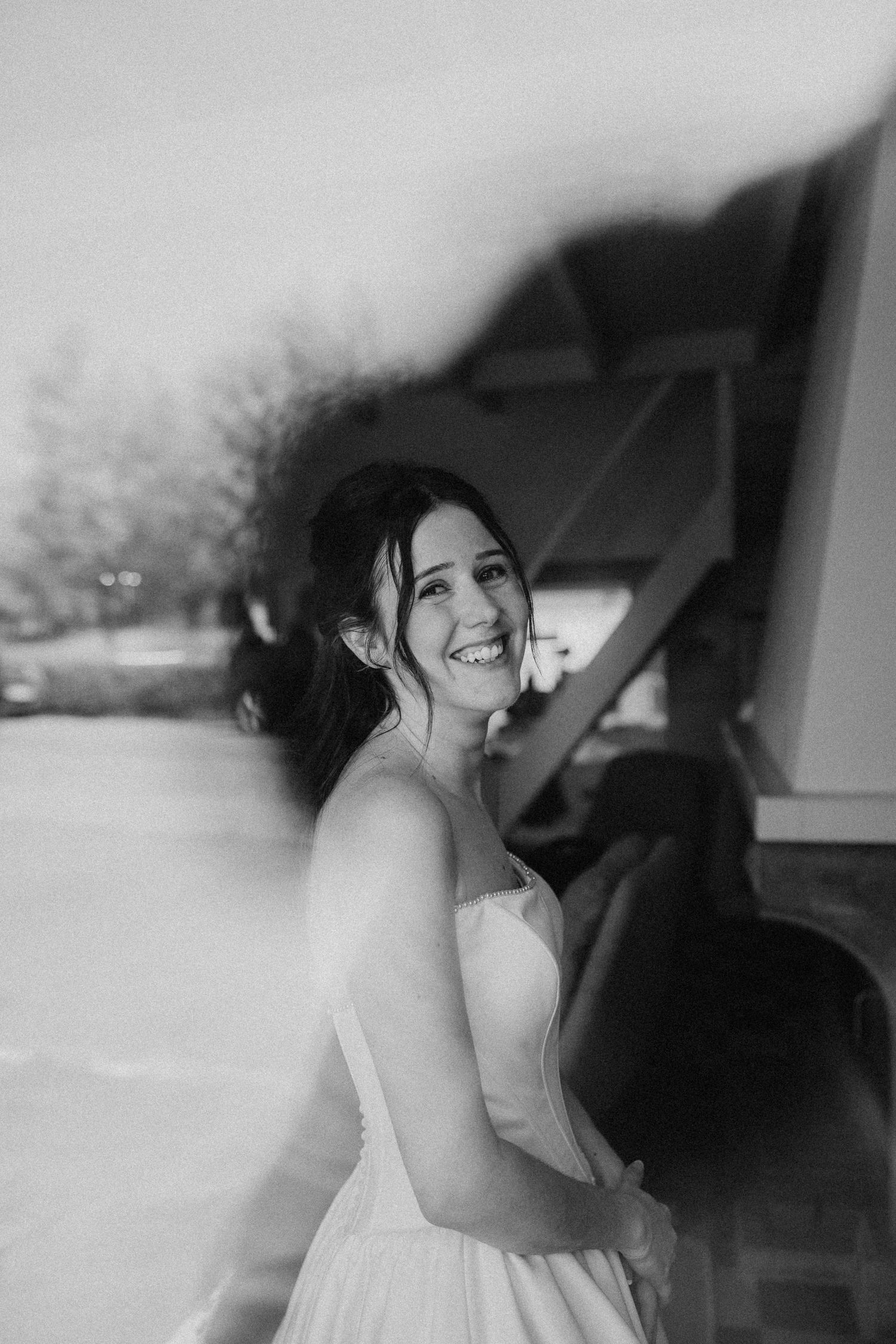 Black and white photo of a smiling bride in a white gown, laughing joyfully outdoors near a rustic building.