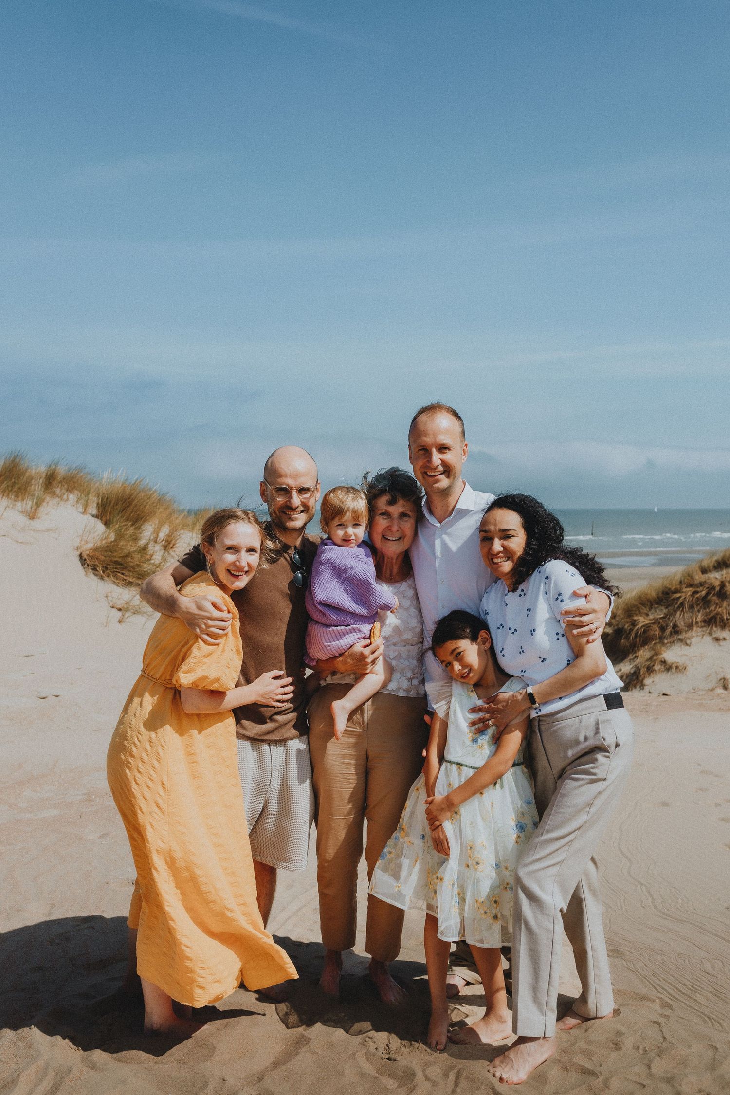 Happy multigenerational family of seven posing together on a sunny sandy beach with dunes.