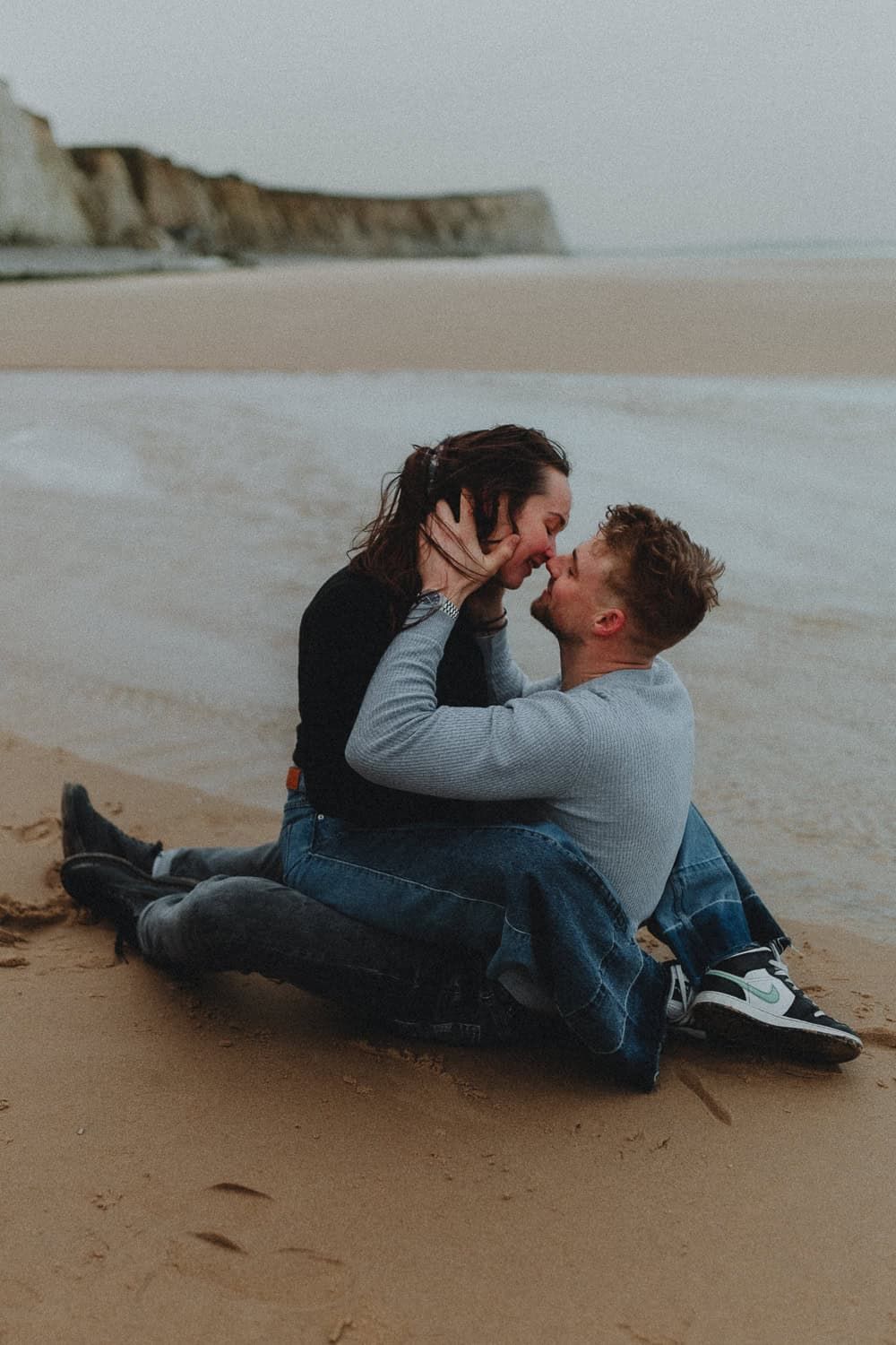 Couple sharing a romantic kiss while sitting on a sandy beach with white cliffs in the background.