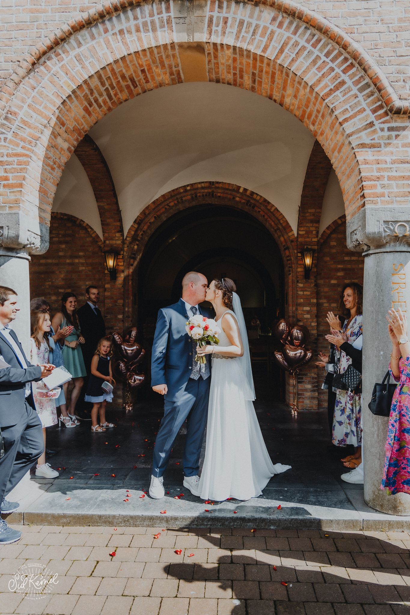 een zonet pas getrouwde koppel die aan de uitgang van de kerk elkaar kussen omring door familie en vrienden op een zonnige dag in Nieuwpoort