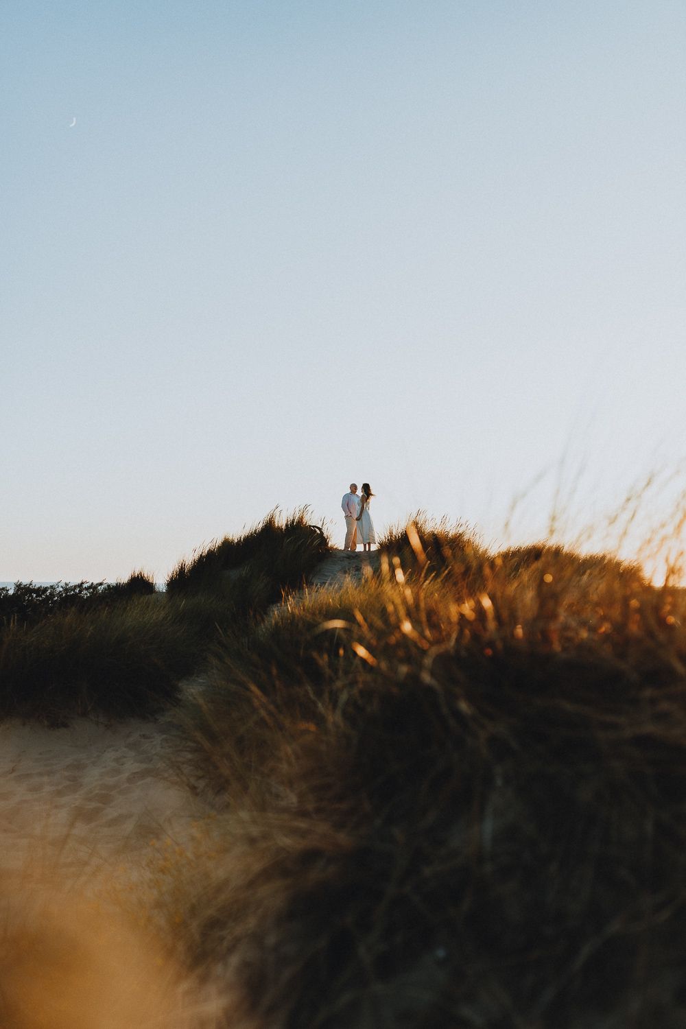 Een koppel staat dicht op een zandduin met gras, onder een blauwe lucht met een sikkelmaan. Rustige en romantische sfeer.
