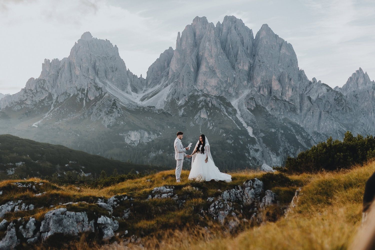 Bride and groom hold hands on a rocky mountain meadow with dramatic Dolomites peaks in the background.