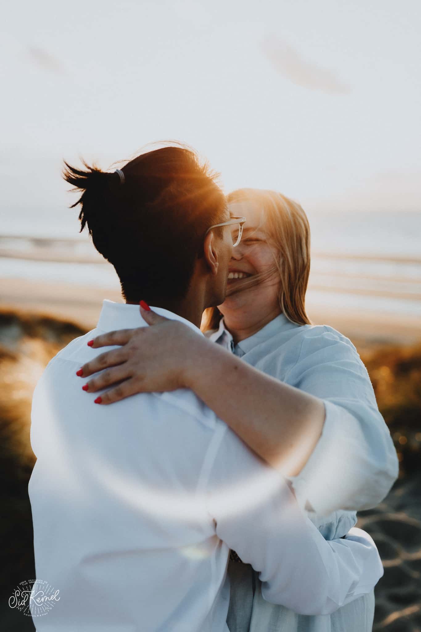 Een koppel omhelst elkaar op een zonovergoten strand in Cadzand. De vrouw heeft roodgelakte nagels en ze glimlachen liefdevol en gelukkig.