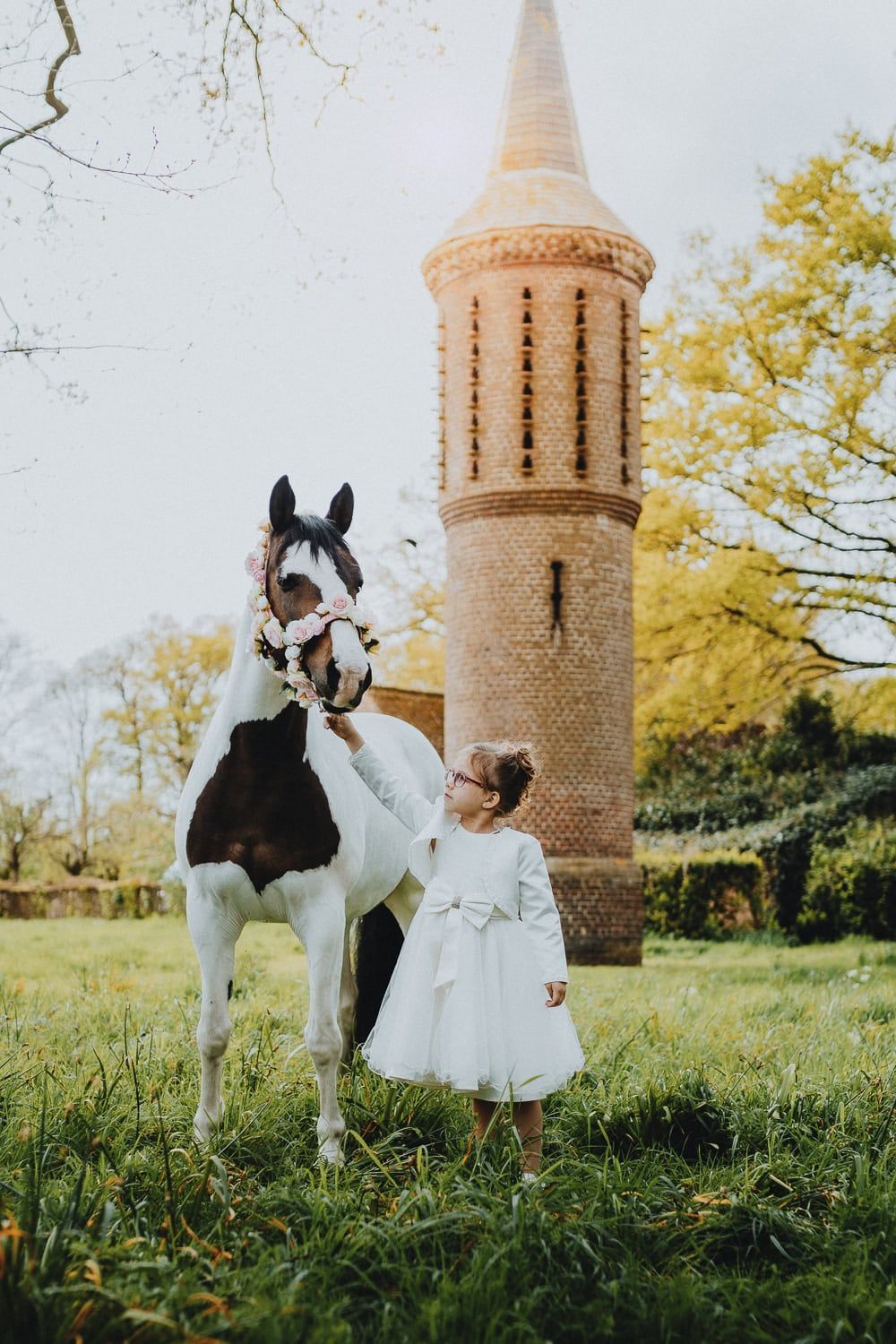 Een meisje in een witte jurk aait zacht een paard met bloemenkrans. Ze staan op gras bij een bakstenen toren onder een heldere lucht.