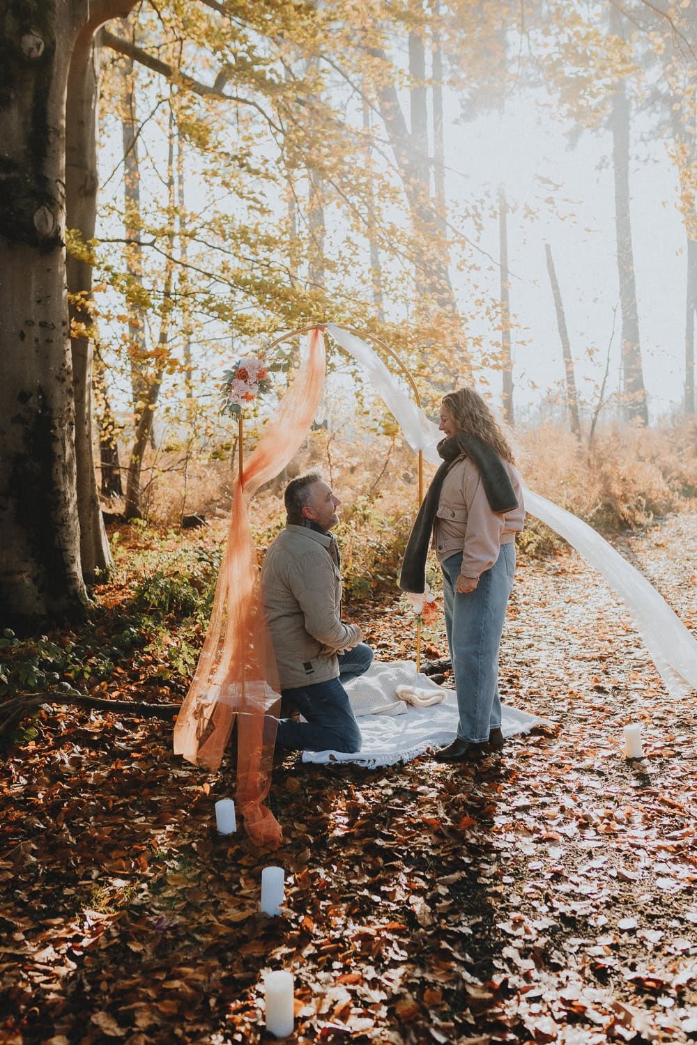 Man proposing to woman in autumn forest with floral arch, candles, and golden foliage.