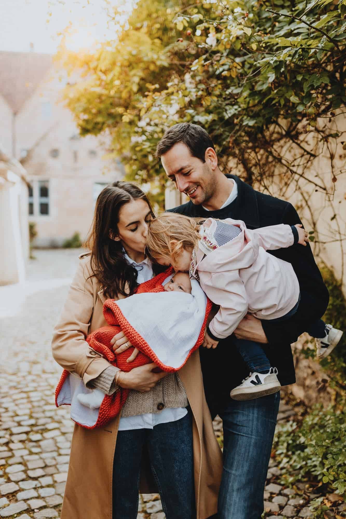 Een gezin van vier lacht liefdevol in Kortrijk. Ouders houden een pasgeborene in een rode deken vast, terwijl hun dochter toekijkt.