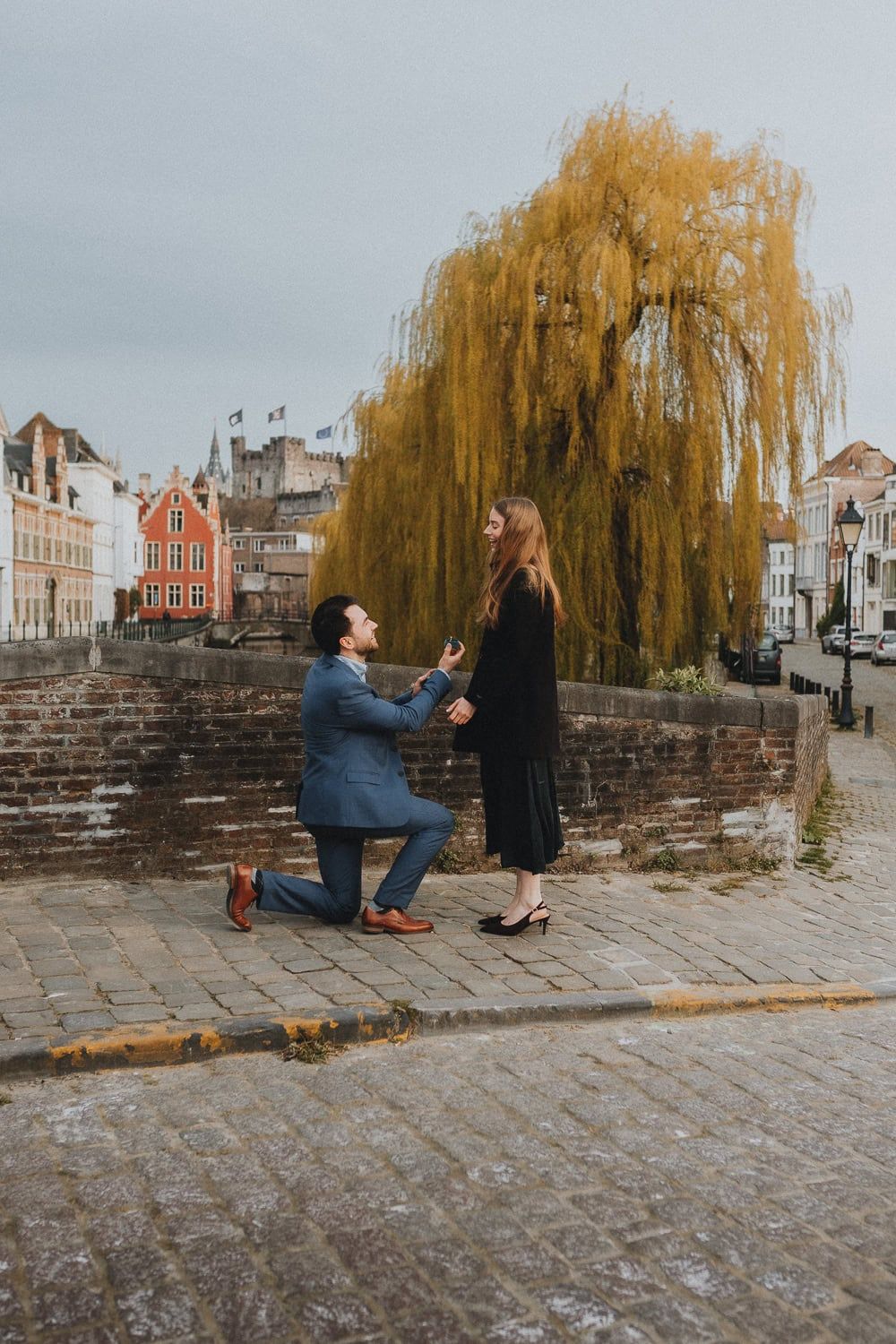 Man proposes on one knee to woman on cobblestone street in Bruges, Belgium, with willow tree and historic buildings backdrop.