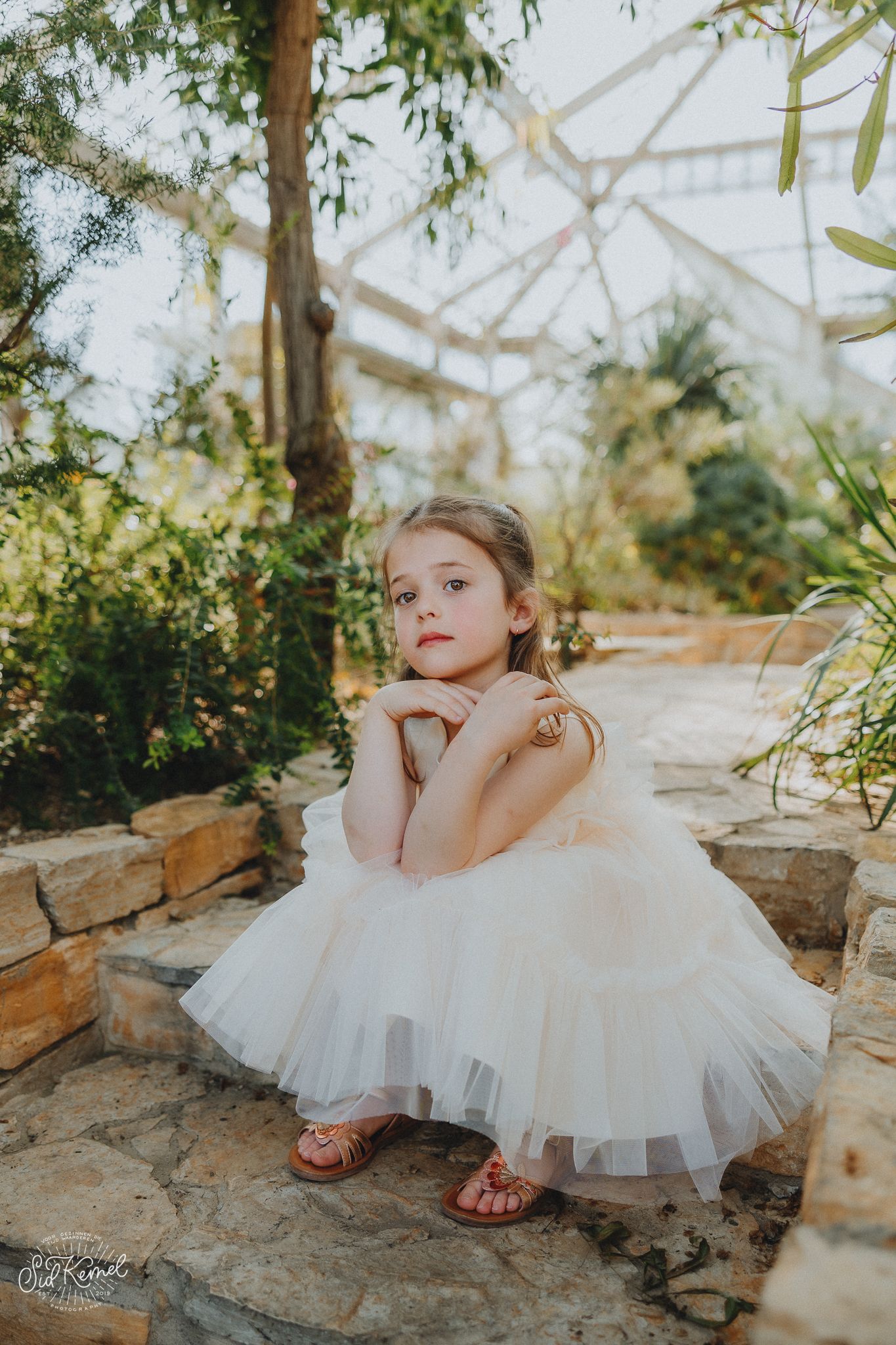 A young girl in a white tulle dress poses gracefully on stone steps in a lush greenhouse garden setting.