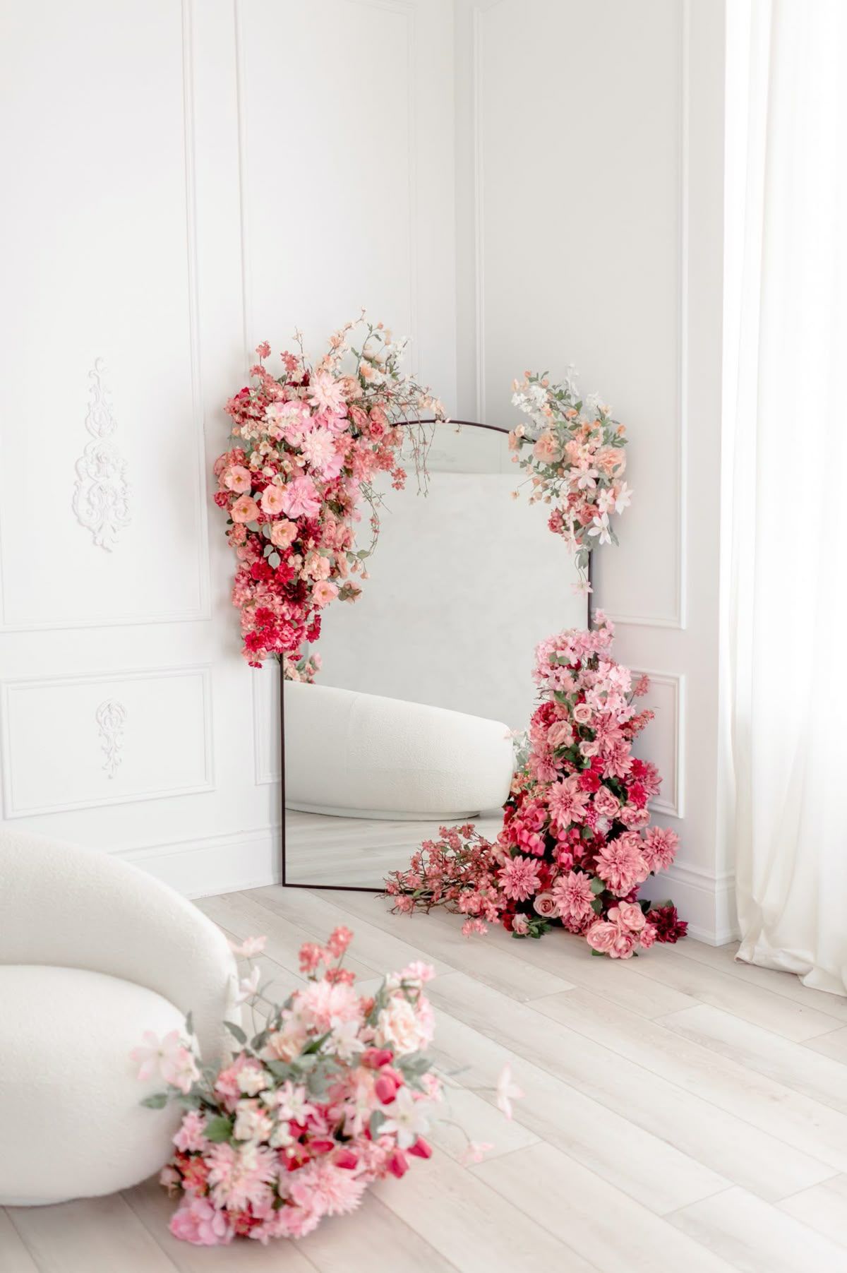 Large mirror decorated with pink and red flowers leaning against white paneled wall in elegant room.