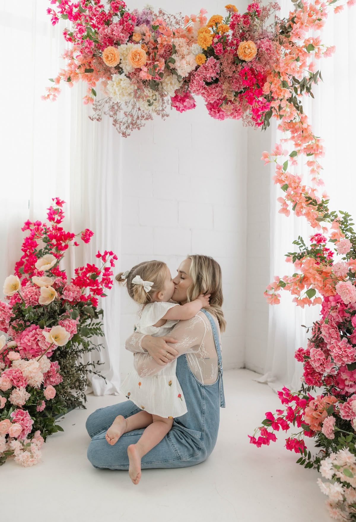 Woman sitting on floor kissing young girl amid lush pink and peach floral arch in white studio.