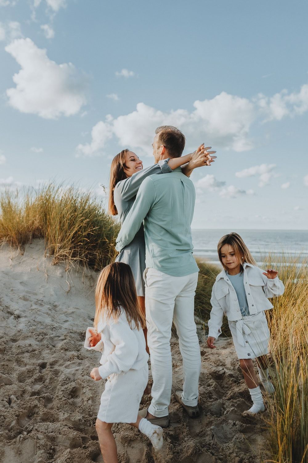 Een gezin van vier geniet van een zonnige dag aan het strand. Ouders omhelzen elkaar bij grasduinen, kinderen spelen dichtbij. Blauwe lucht.