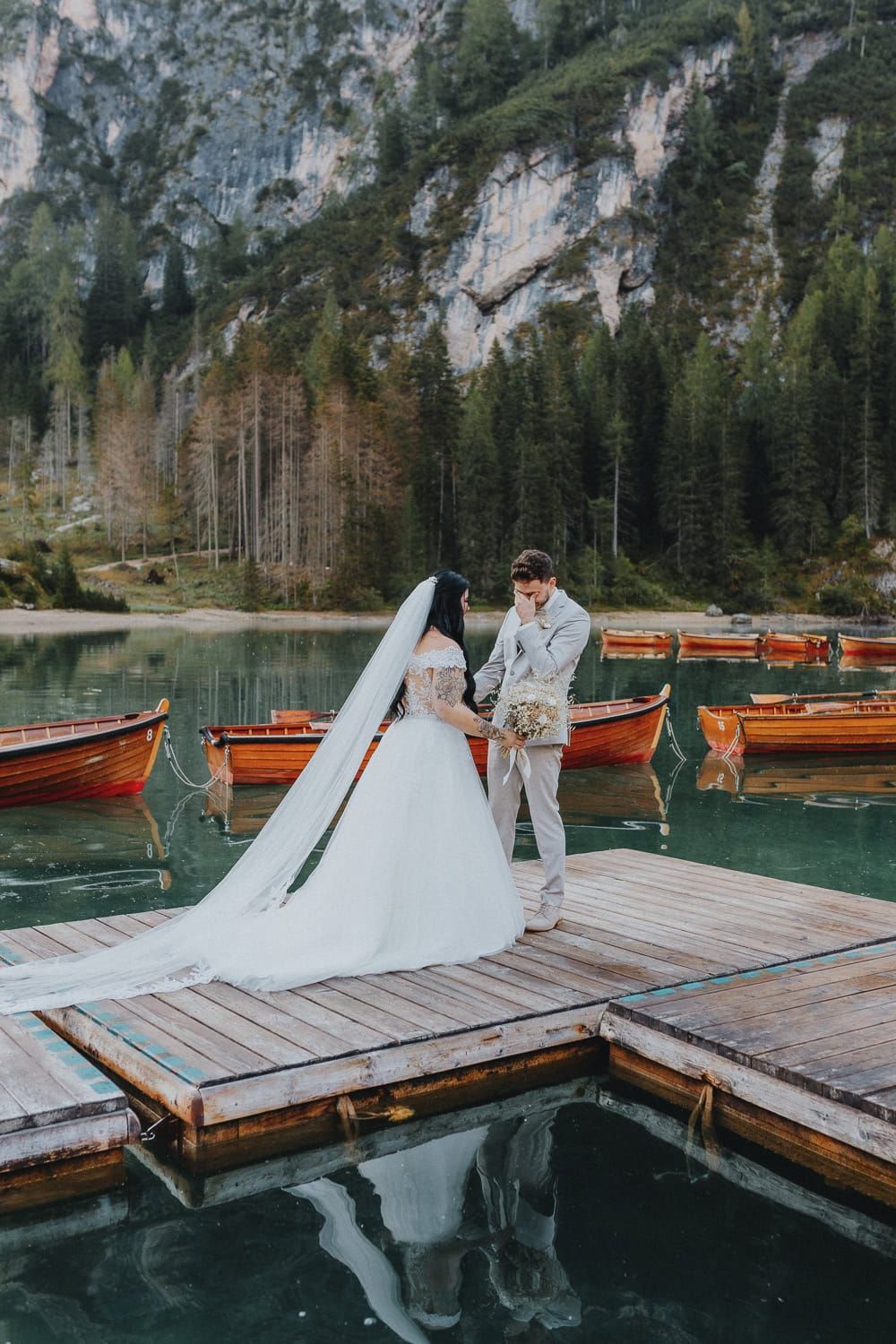 Wedding couple holding hands on wooden dock at alpine lake with red boats and mountain backdrop.