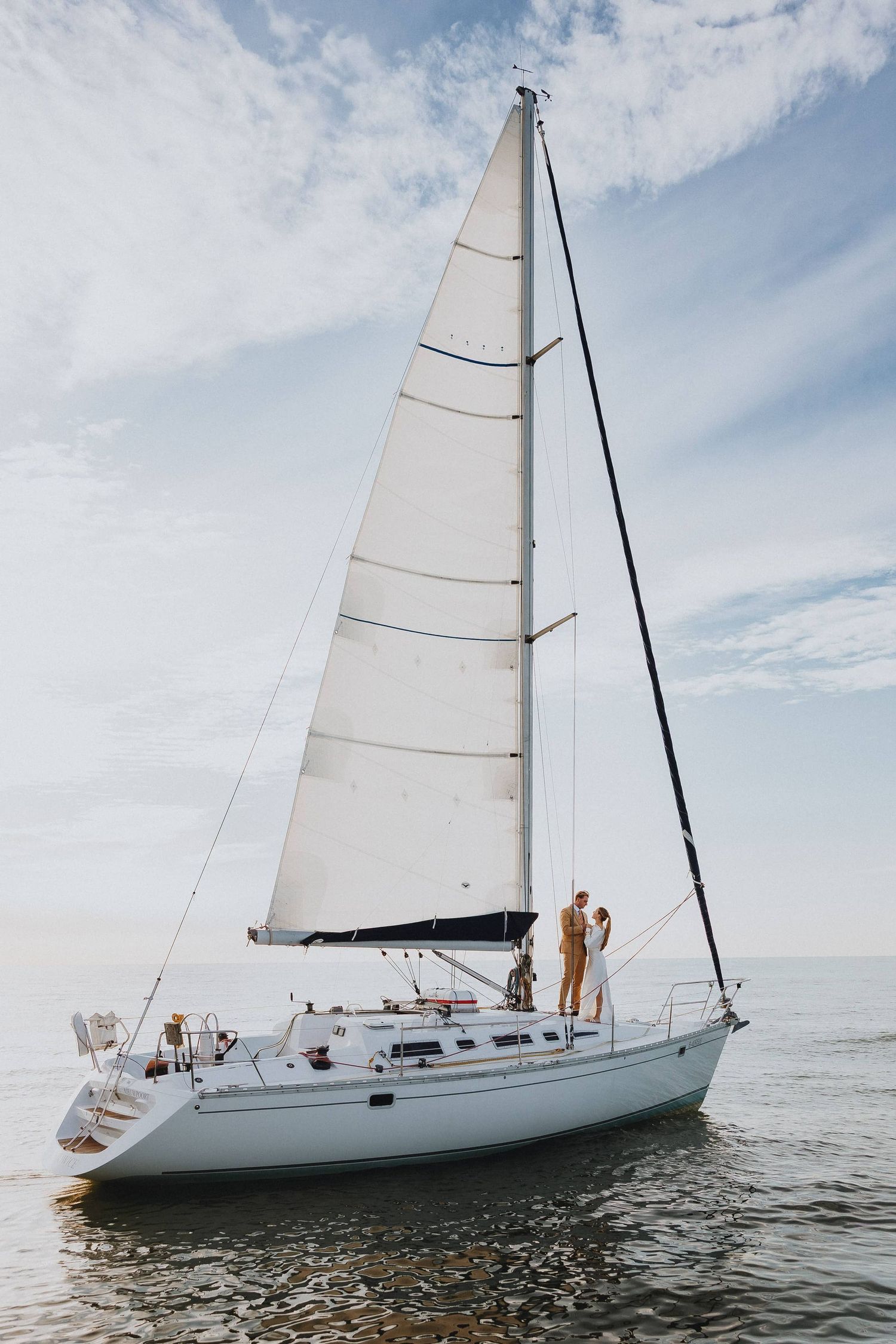 Couple embracing on a white sailboat with full sails on calm open water under a partly cloudy sky.