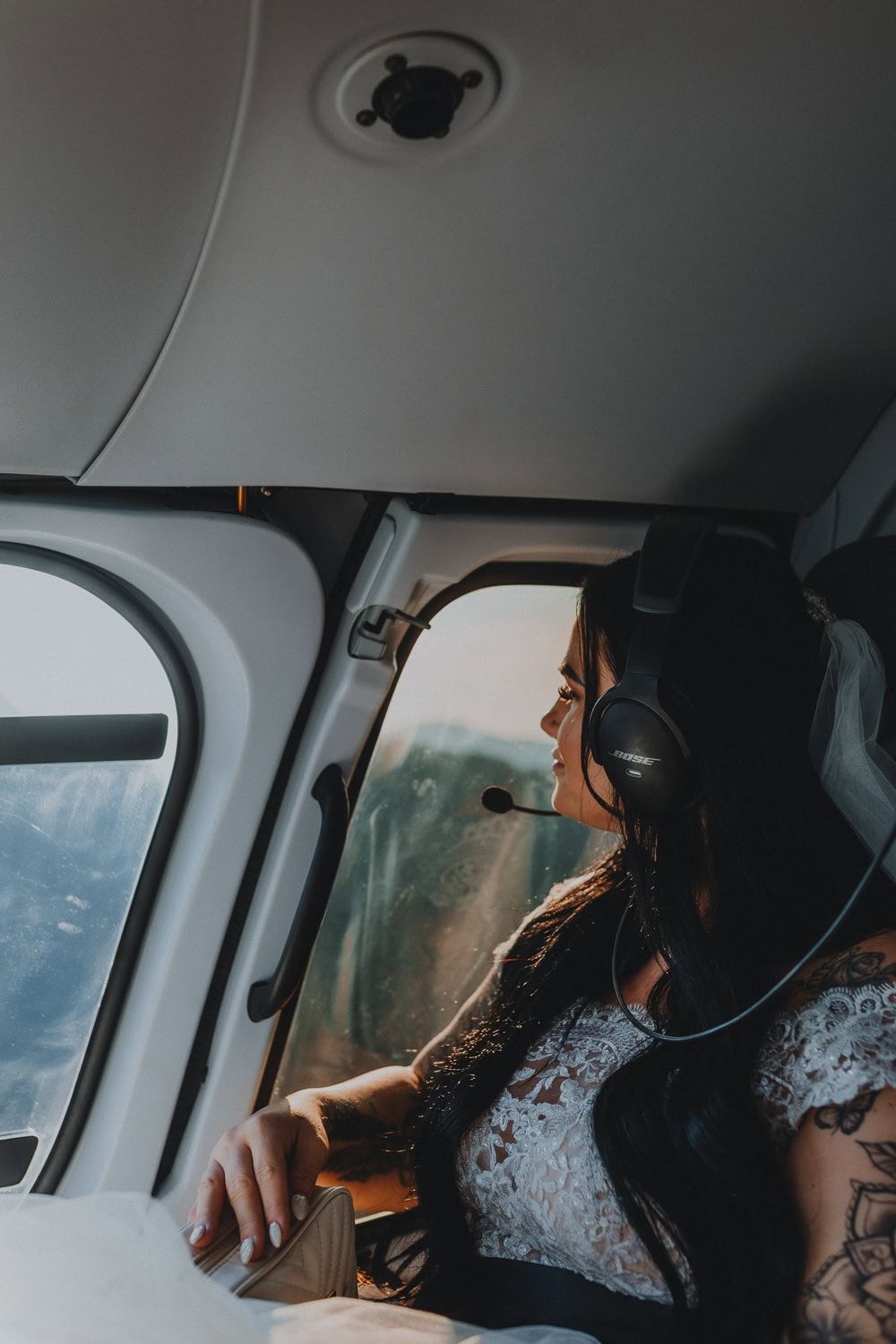 Bride in lace wedding dress wearing aviation headset looking out helicopter window at mountain views.