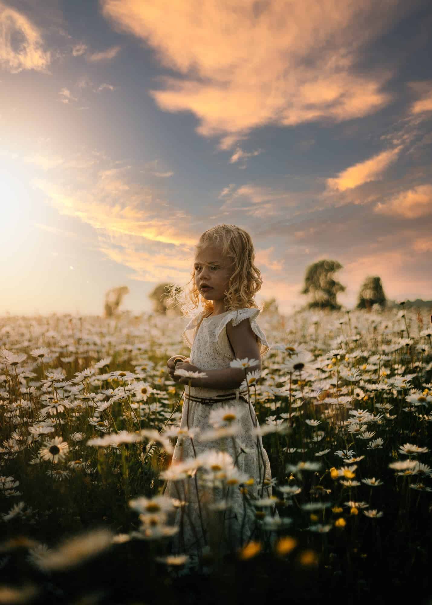 Een meisje in een witte jurk staat in een madelievenveld bij zonsondergang. De lucht kleurt warm oranje en roze, een sereen tafereel.