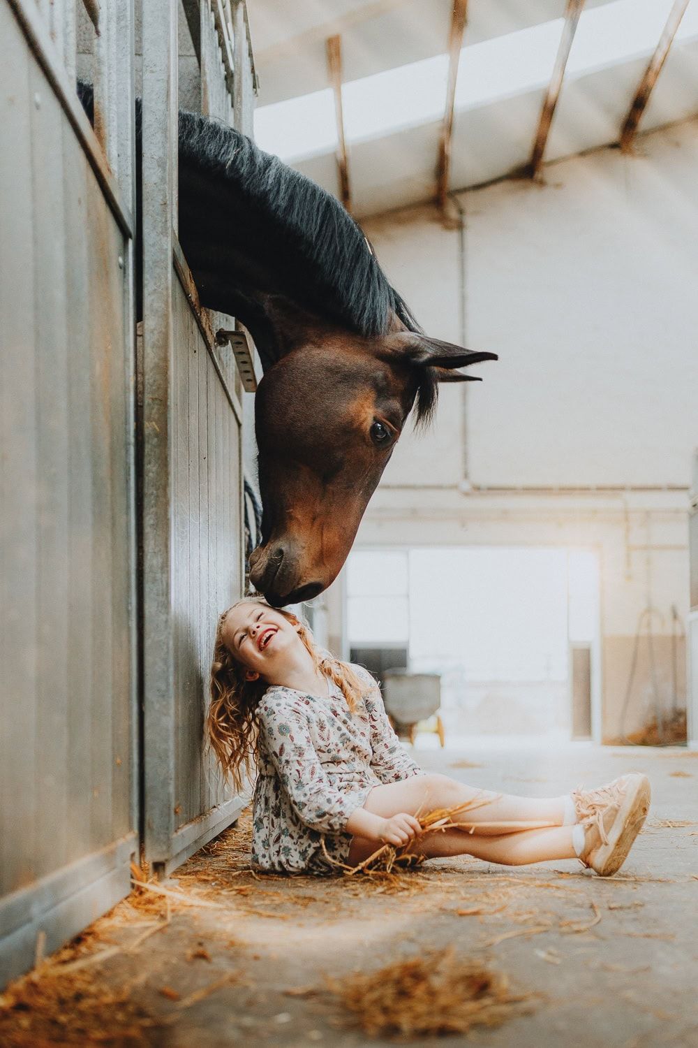 Een meisje lacht op de stalvloer terwijl een paard haar zachtjes aanraakt. Zacht zonlicht zorgt voor een warme en tedere ambiance.