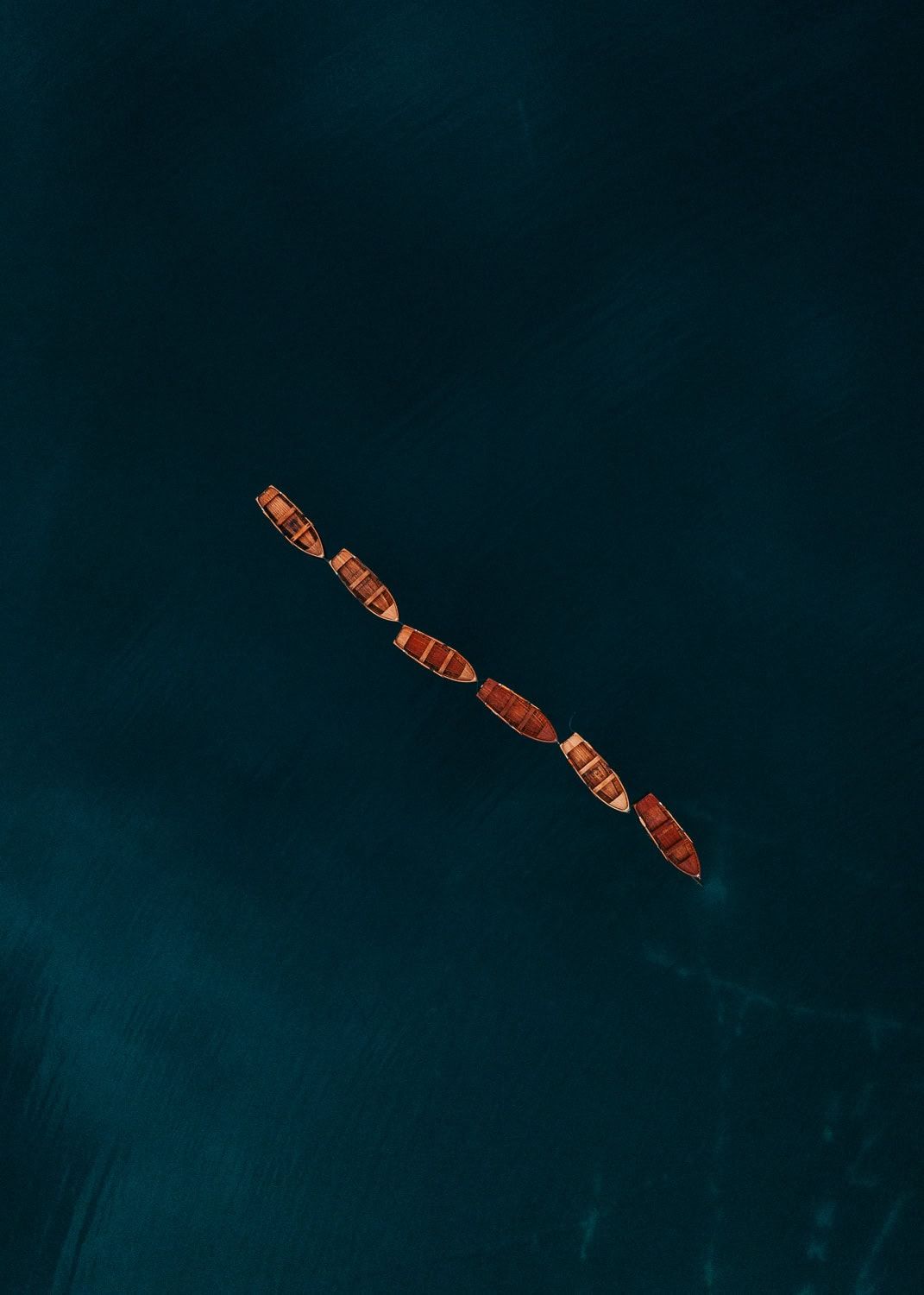Aerial top-down view of wooden boats lined up diagonally on deep teal ocean water.