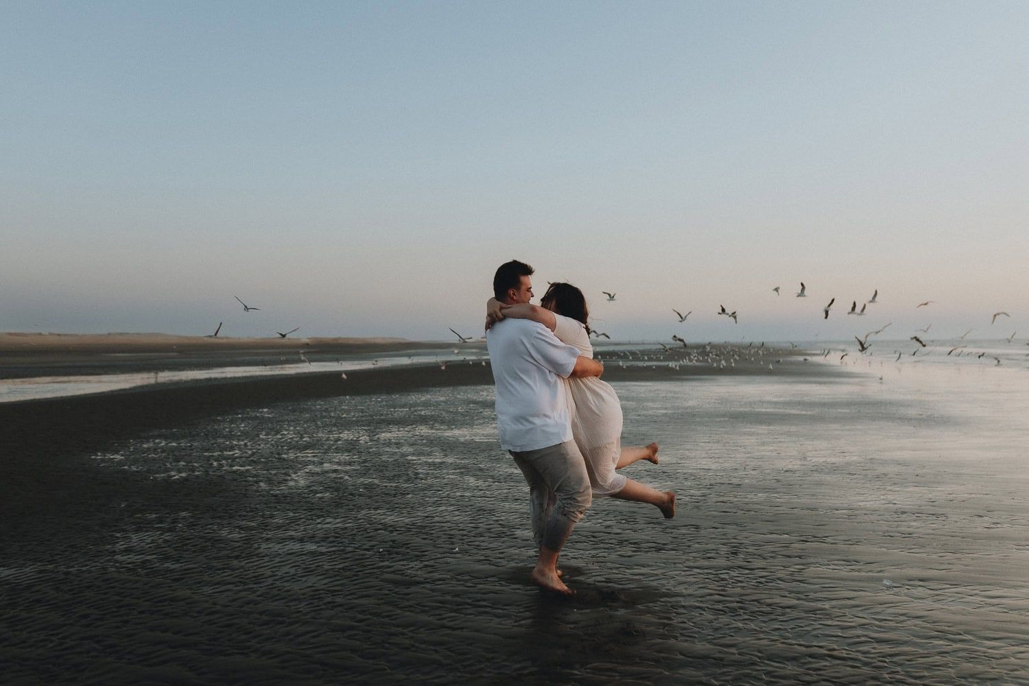 Couple embracing on beach at dusk with birds flying overhead and gentle waves at their feet.