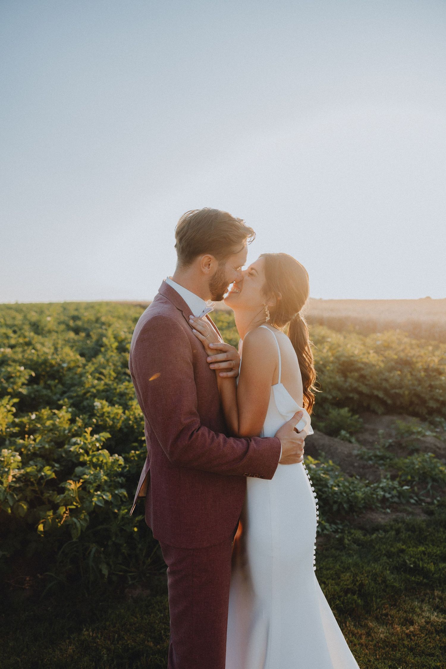 A couple in wedding attire shares a romantic moment at sunset in a lush green field.