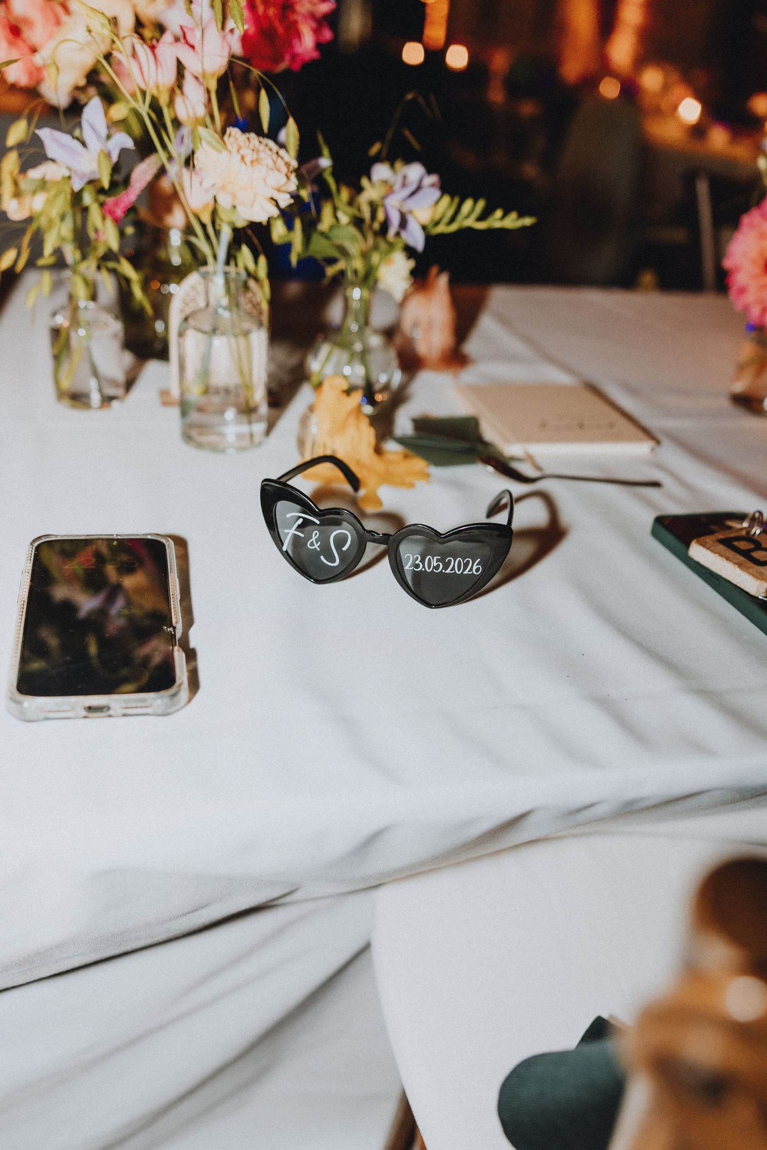 Heart-shaped sunglasses with wedding date 13.05.2026 and initials on white table with floral centerpiece.