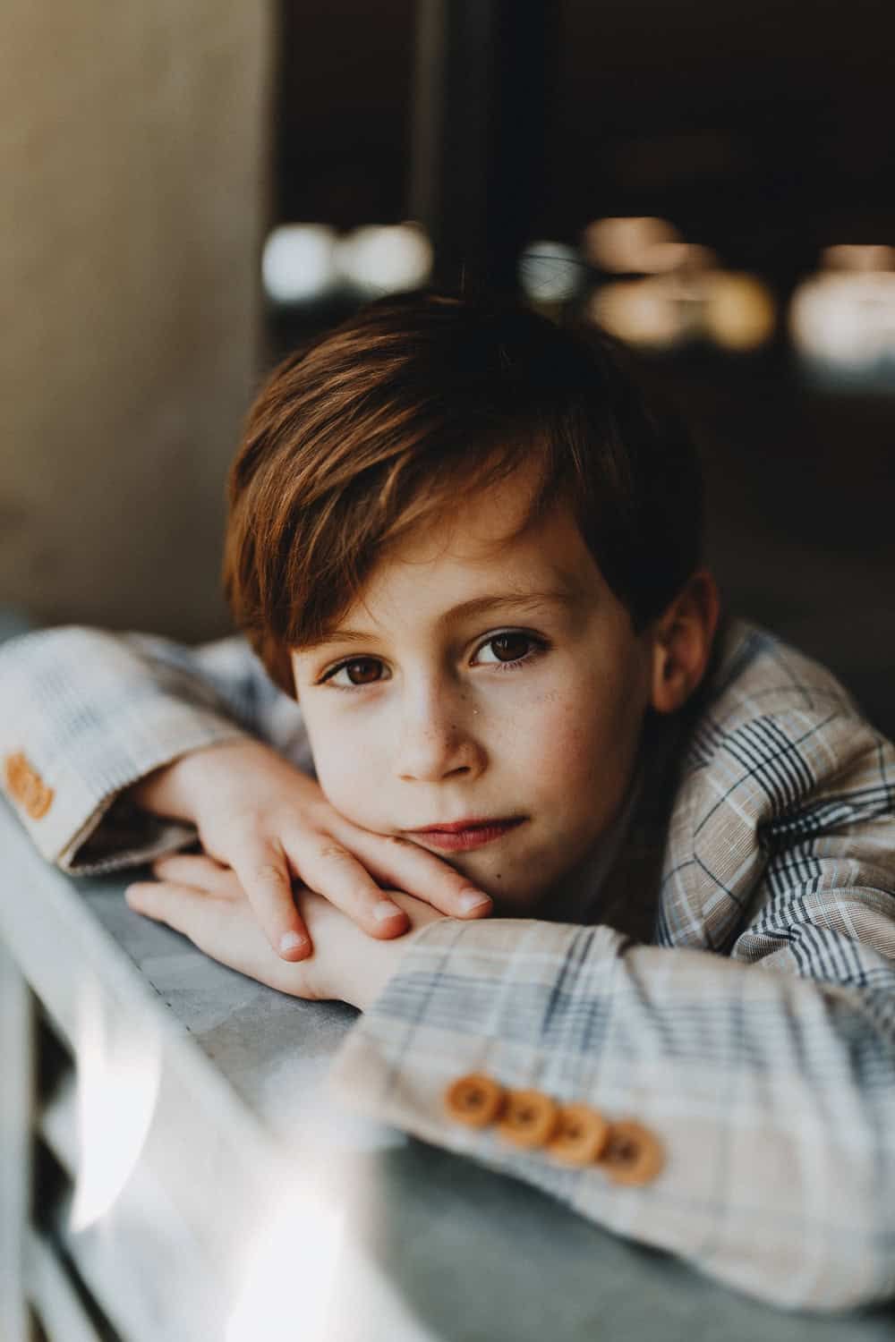 Een jongen met kastanjebruin haar leunt met gekruiste armen en draagt een geruit jasje. Hij kijkt zacht en bedachtzaam in de camera.